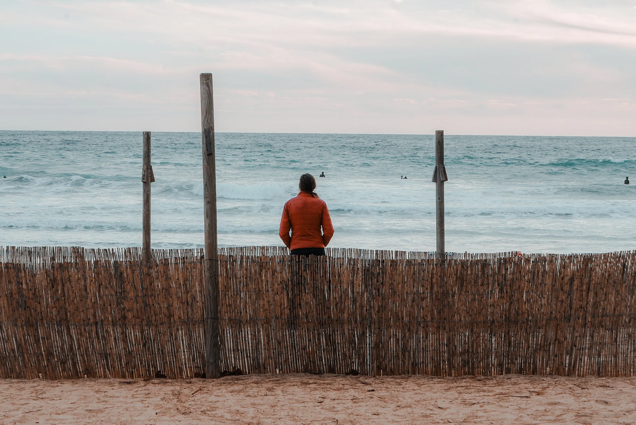 A person in a red jacket stands behind a bamboo fence looking at the ocean, with a few people swimming in the water in the distance.