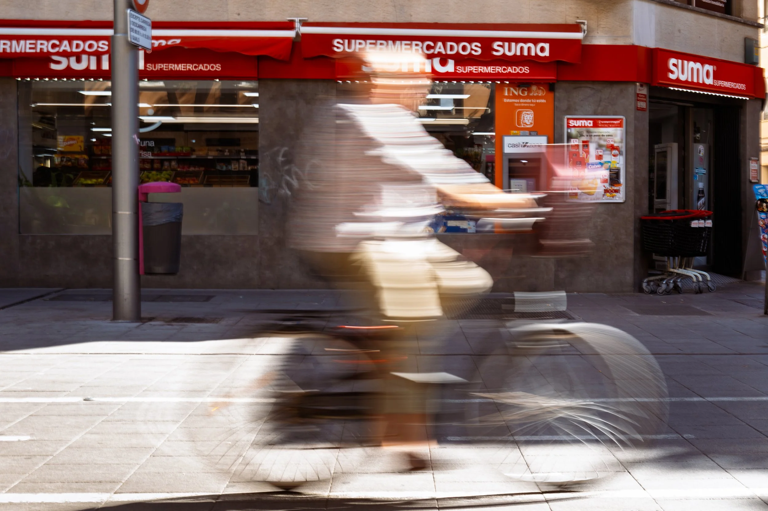 Blurry person riding a bicycle past a grocery store with red signage that reads 'Supermercados Suma' on a city sidewalk.