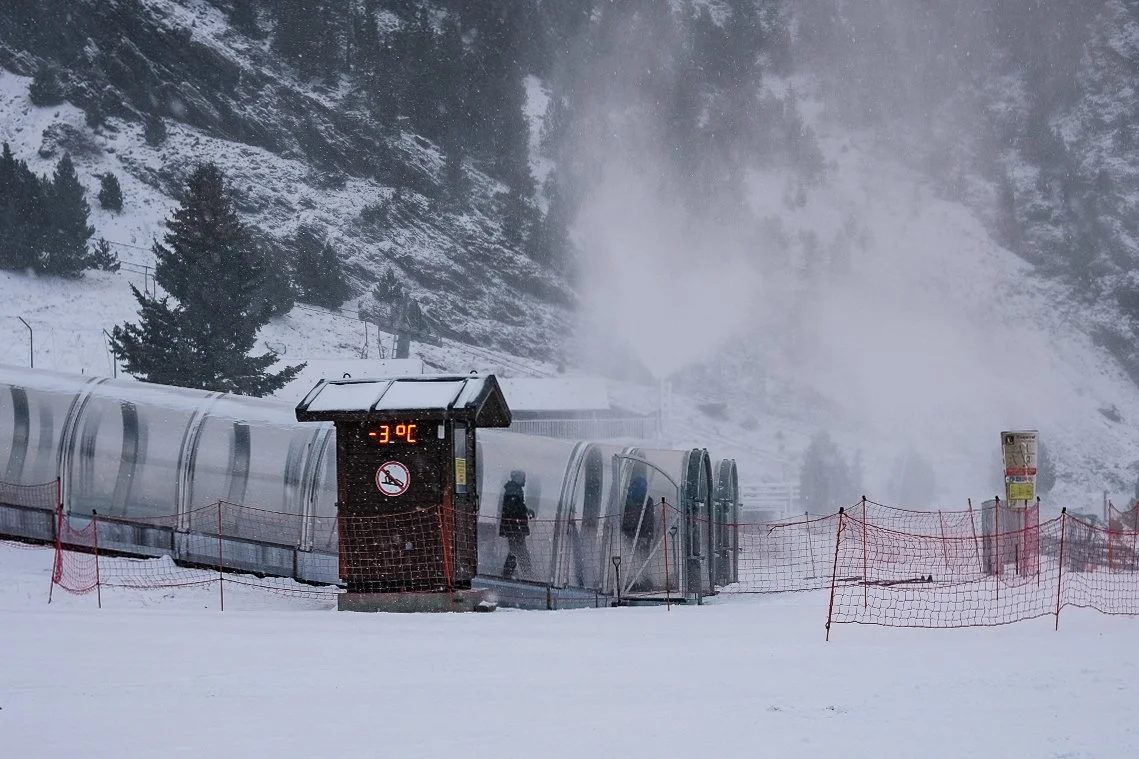Snowy ski resort with temperature display showing -3°C, covered conveyor belt, red safety netting, trees, and snowy mountain backdrop.