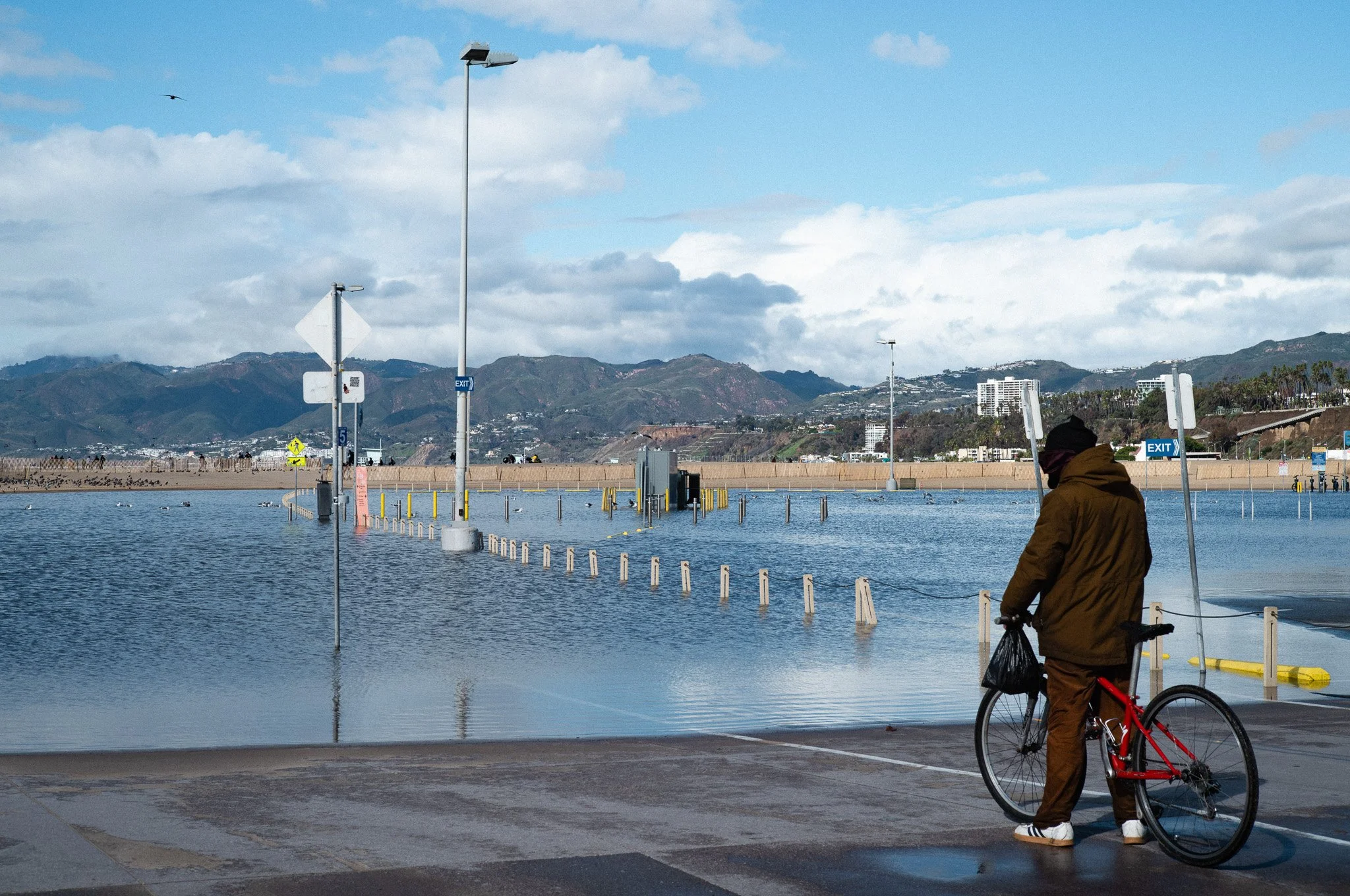 Person in brown jacket and pants holding a red bicycle near a flooded area with water covering the parking lot, and mountains visible in the background under a partly cloudy sky.