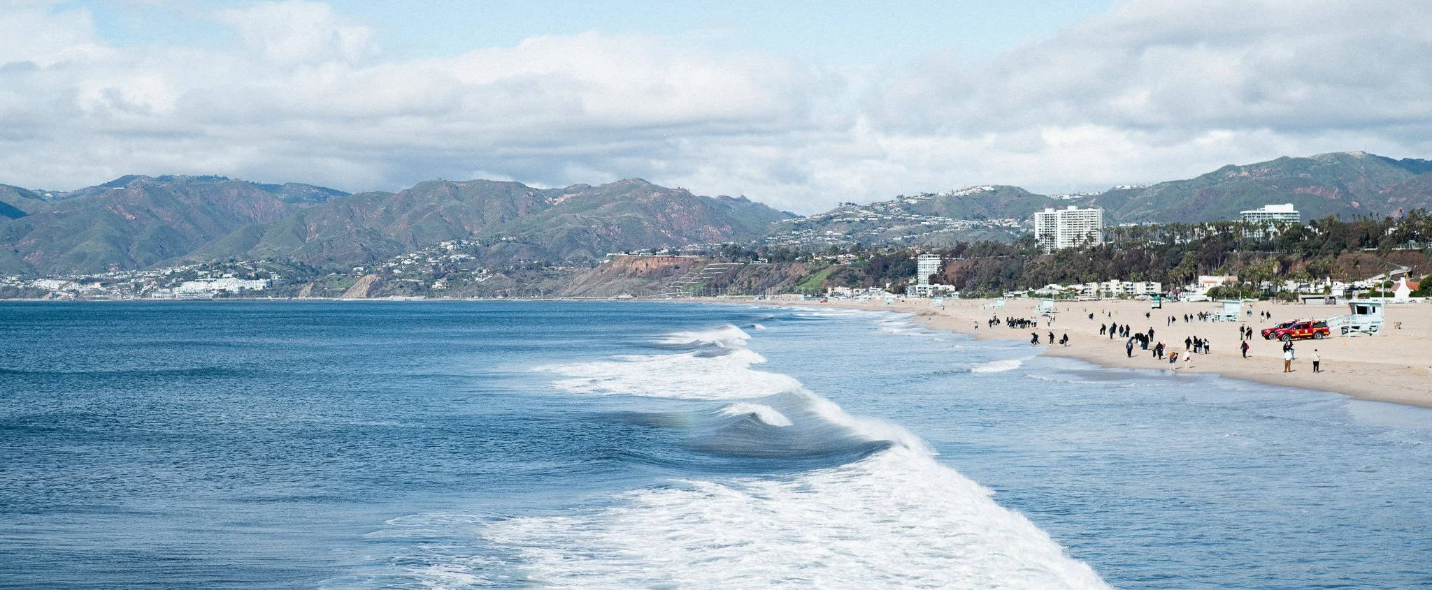 View of a beach with people walking and enjoying the shoreline, ocean waves, and city buildings with hills in the background under a partly cloudy sky.