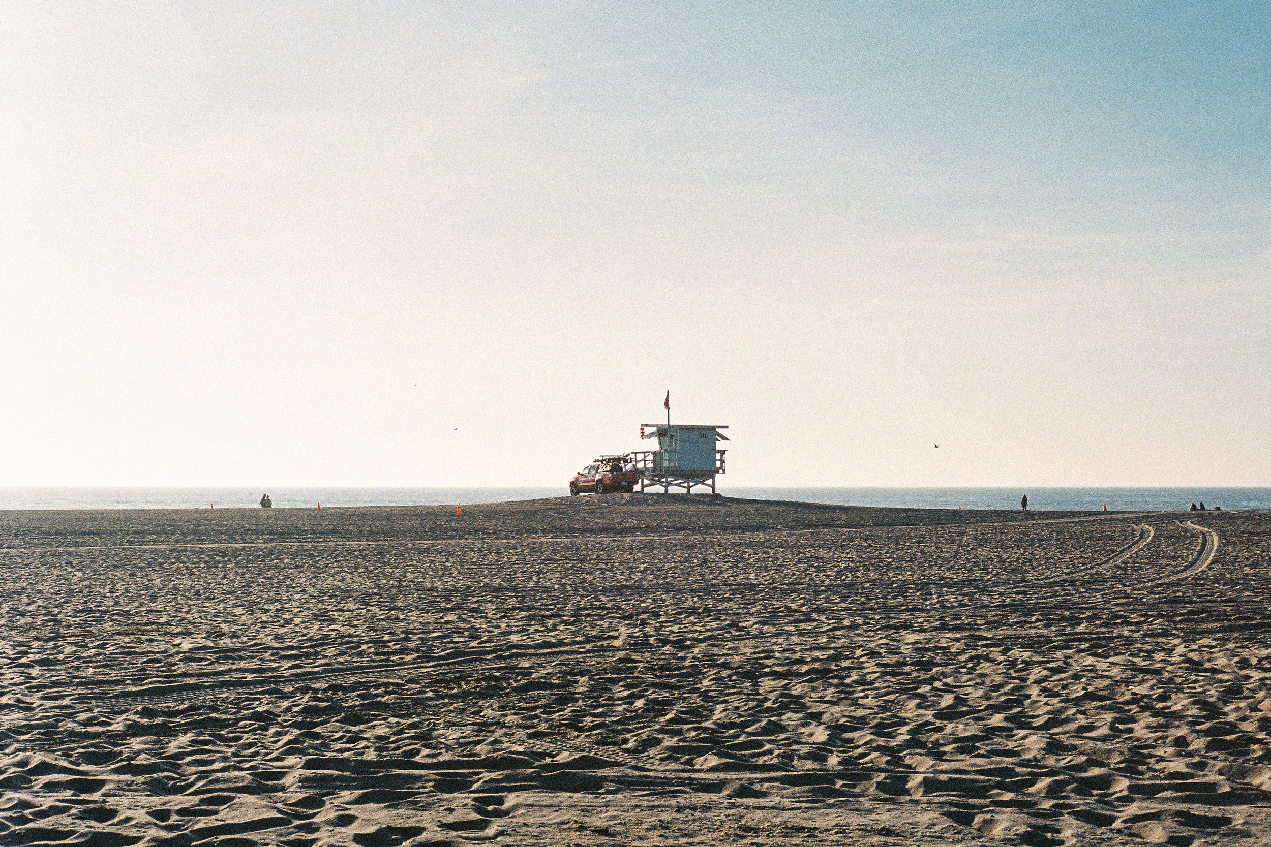 A lifeguard tower and vehicle on a sandy beach with a few people in the distance, ocean and clear sky in the background.