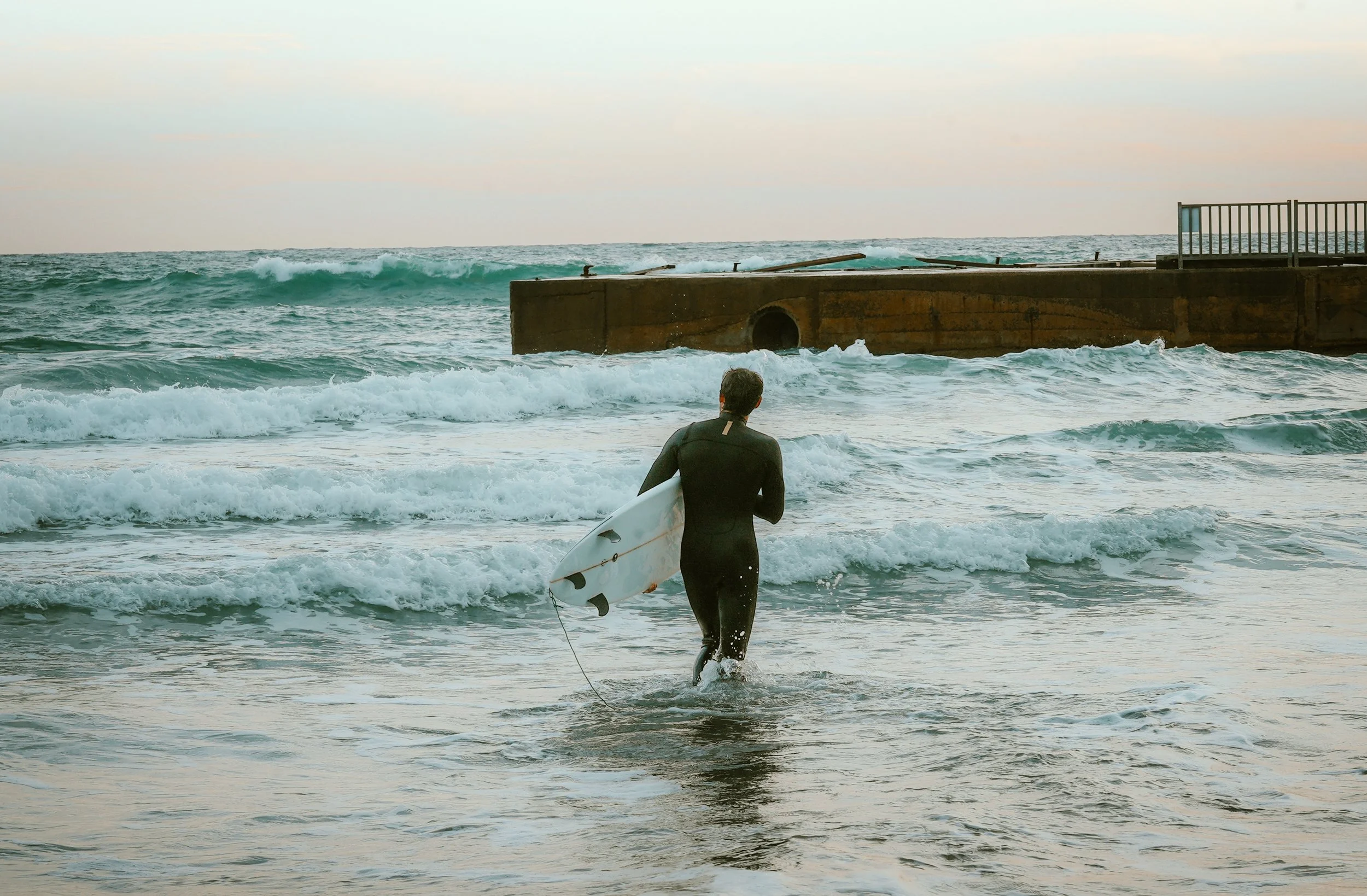 Surfer holding a surfboard walking into the ocean near a concrete pier