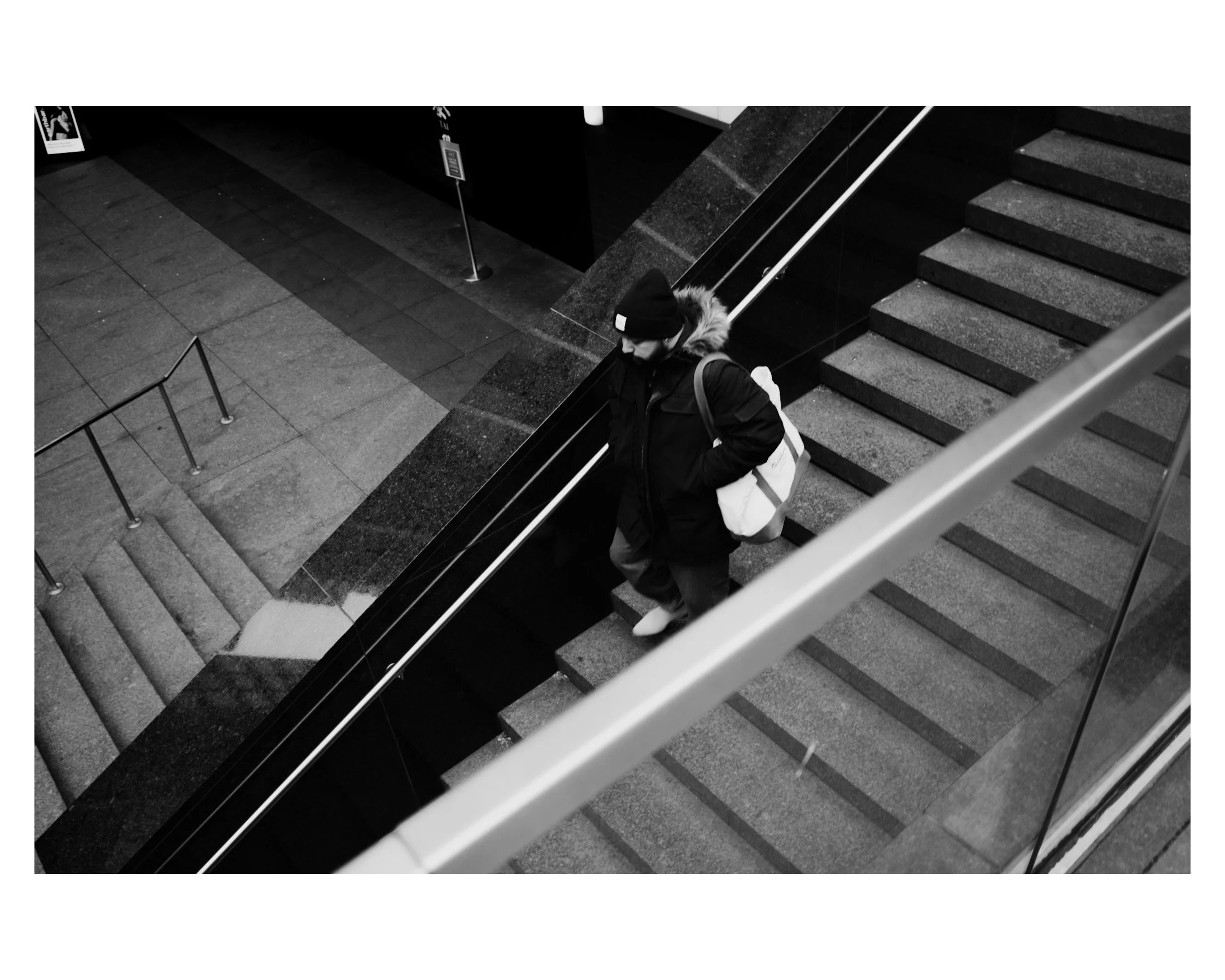 A person wearing a winter coat, beanie, and backpack walking down staircase in a modern indoor building, viewed from above in black and white.