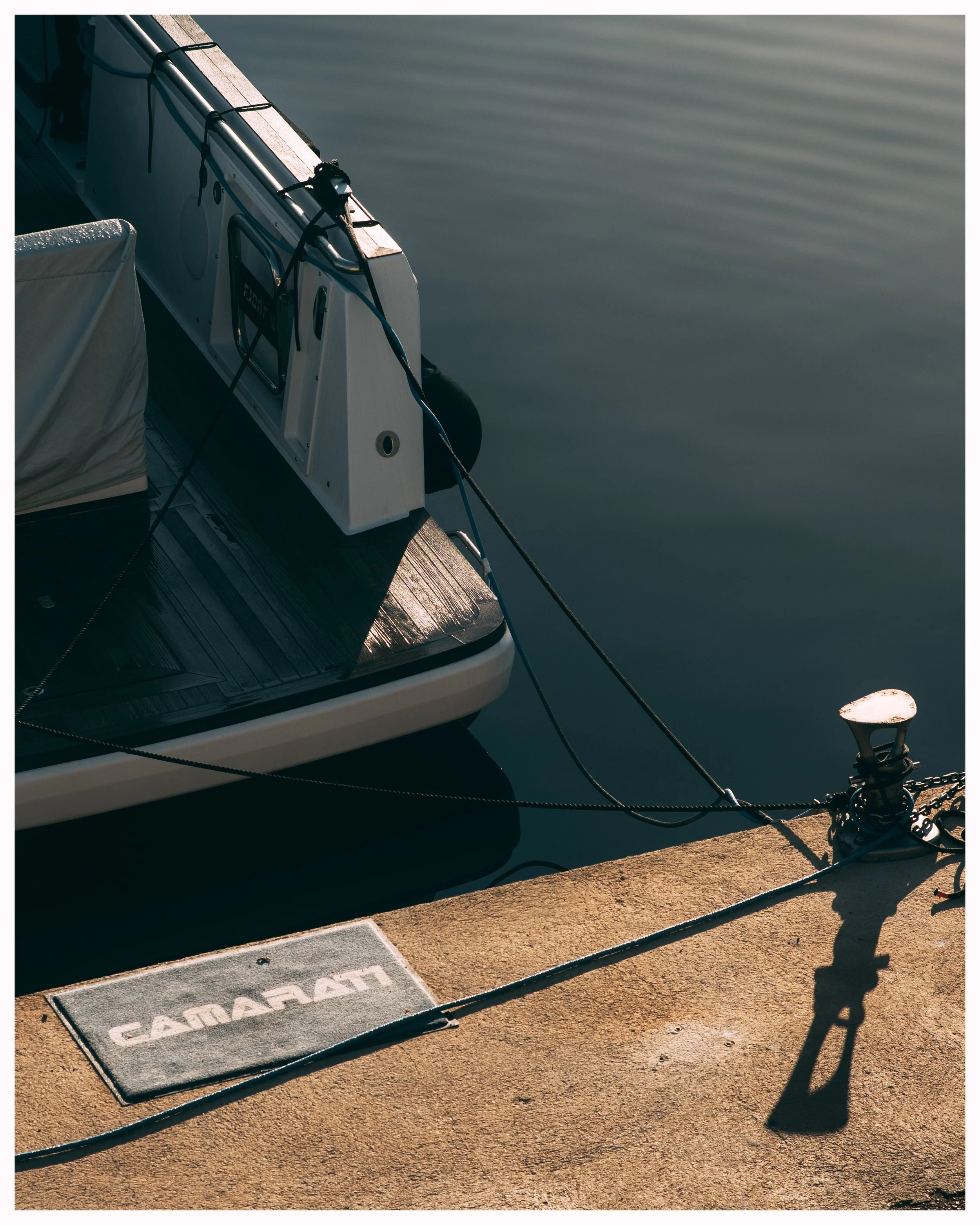 A close-up of a boat docked at a pier, showing the stern with the boat's name in Cyrillic, and the dock's concrete surface with a metal plate and cleat with ropes tied to it.