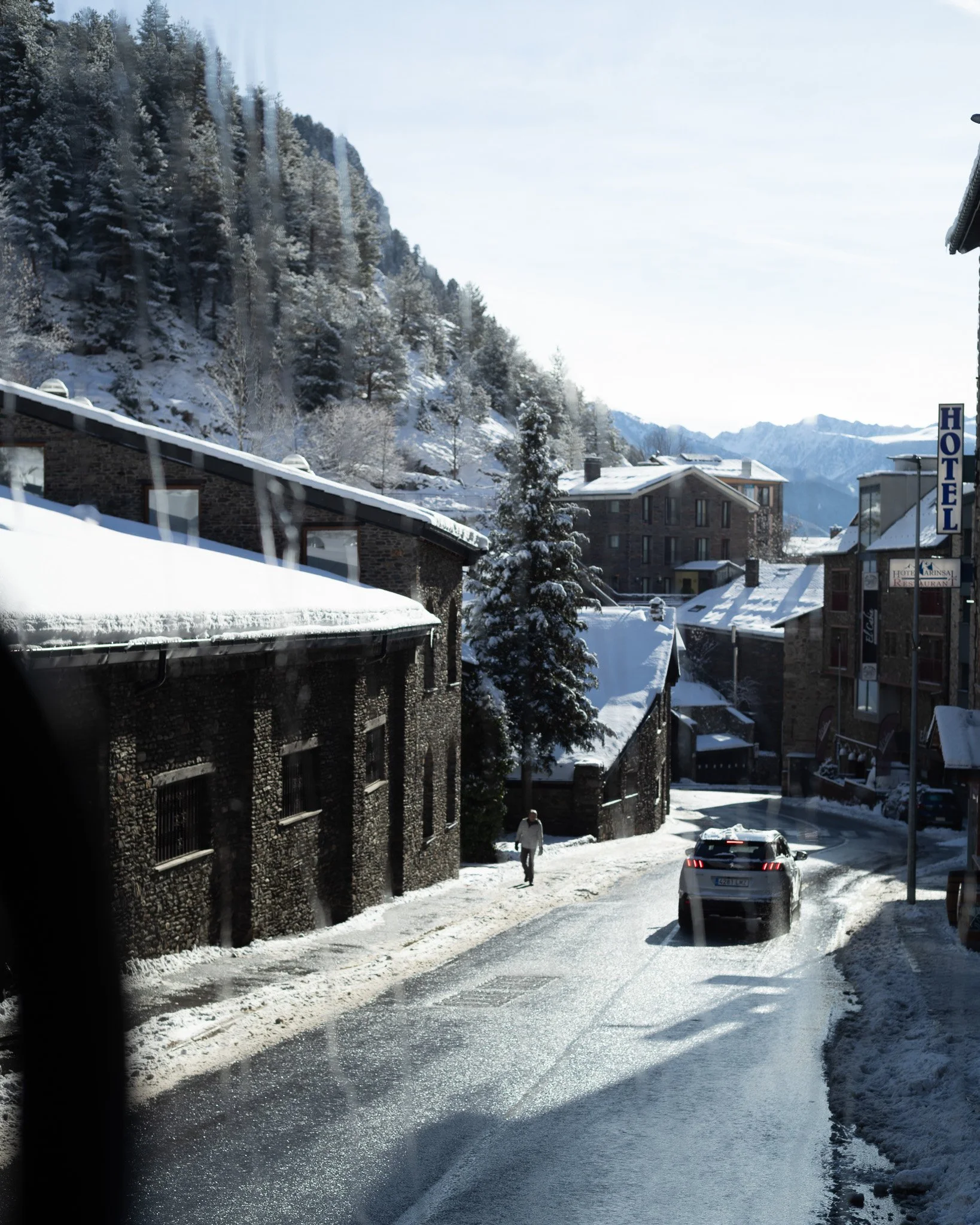 Snow-covered street in a mountainous town with buildings, a hotel sign, and a car driving on the icy road
