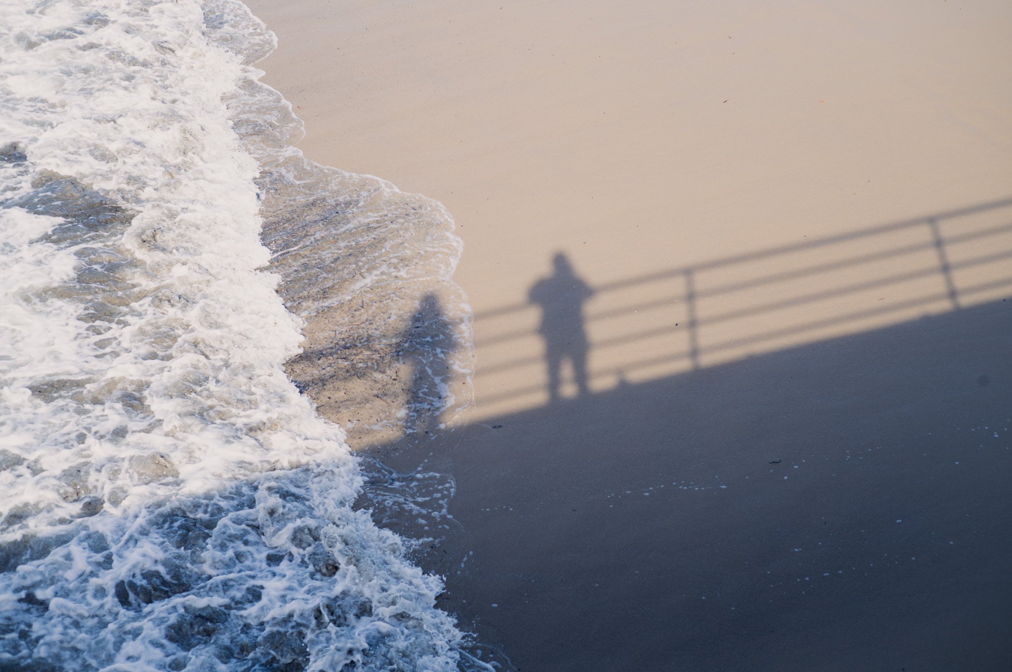 Shadows of two people, one with a camera, cast on the sandy beach near the shoreline, with waves washing up on the sand