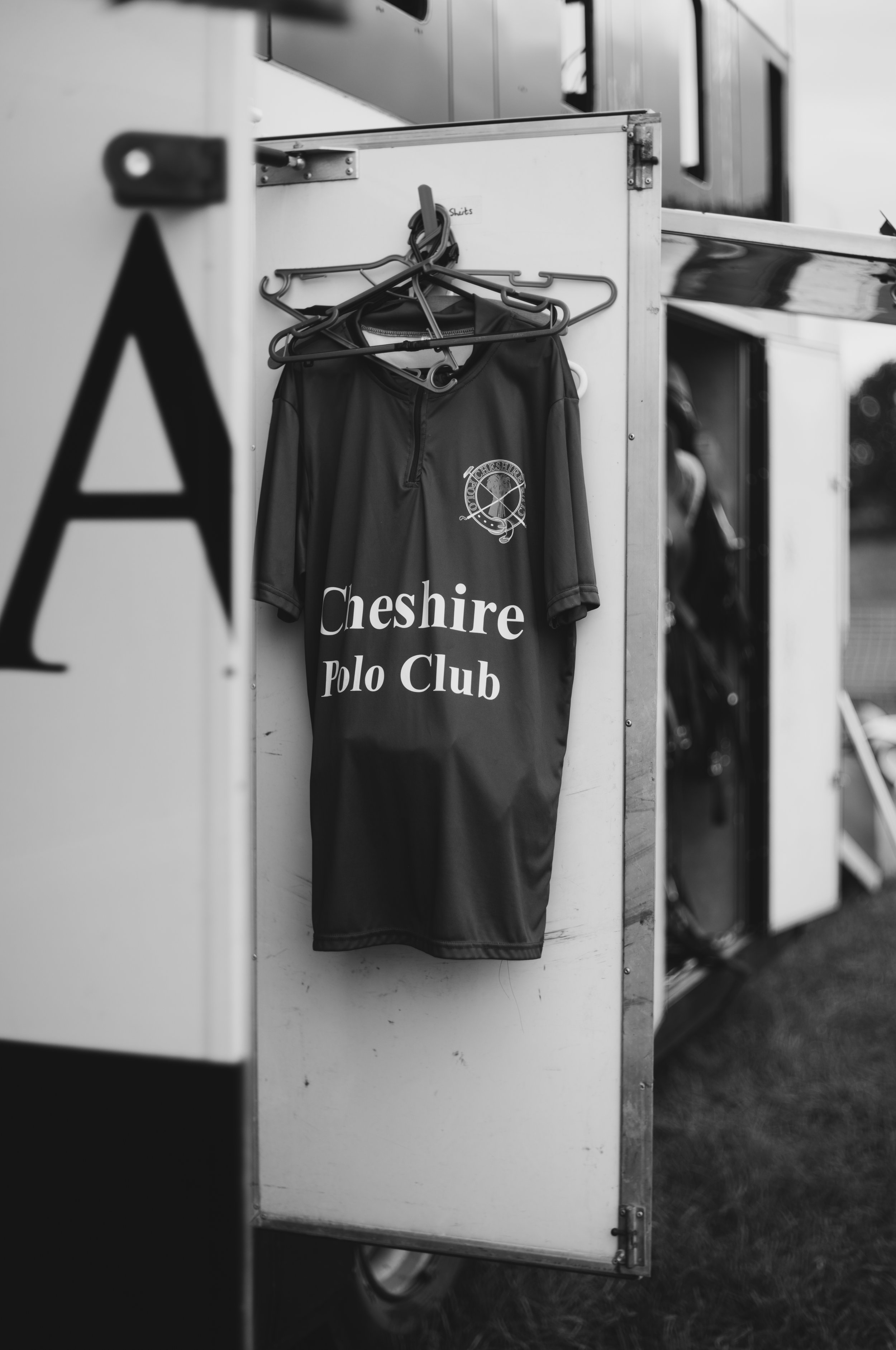 A black and white photo of an open locker with a polo shirt hanging inside. The shirt has the words 'Cheshire Polo Club' printed on it, along with a logo featuring crossed polo mallets and a ball.