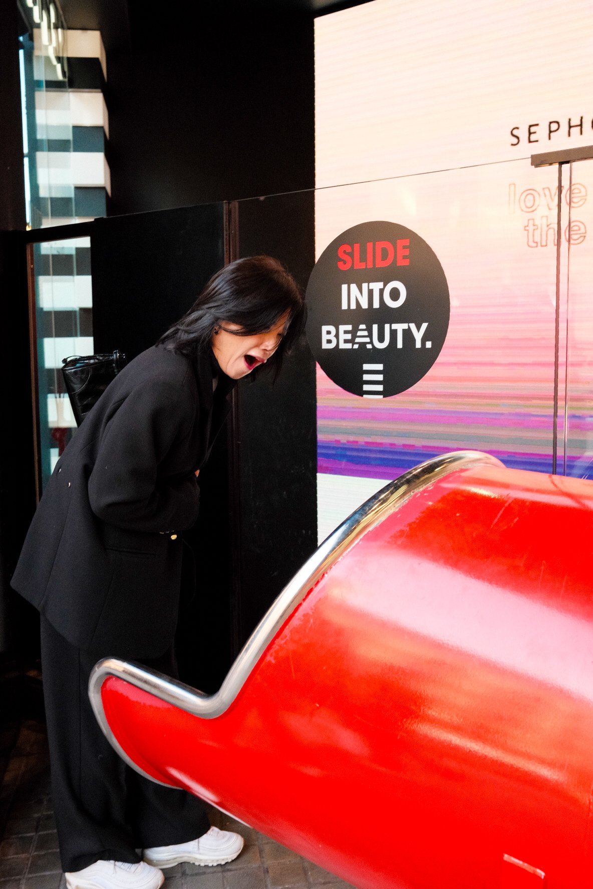 A woman in black clothing leaning over a red, shiny, curved object, looking inside with surprise, in front of a sign that reads 'Slide into Beauty' with colorful background.