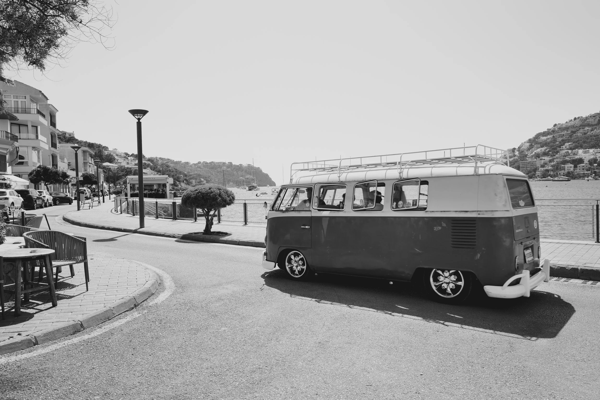 A vintage Volkswagen camper van parked by a waterfront with hills and buildings in the background, black and white photo.
