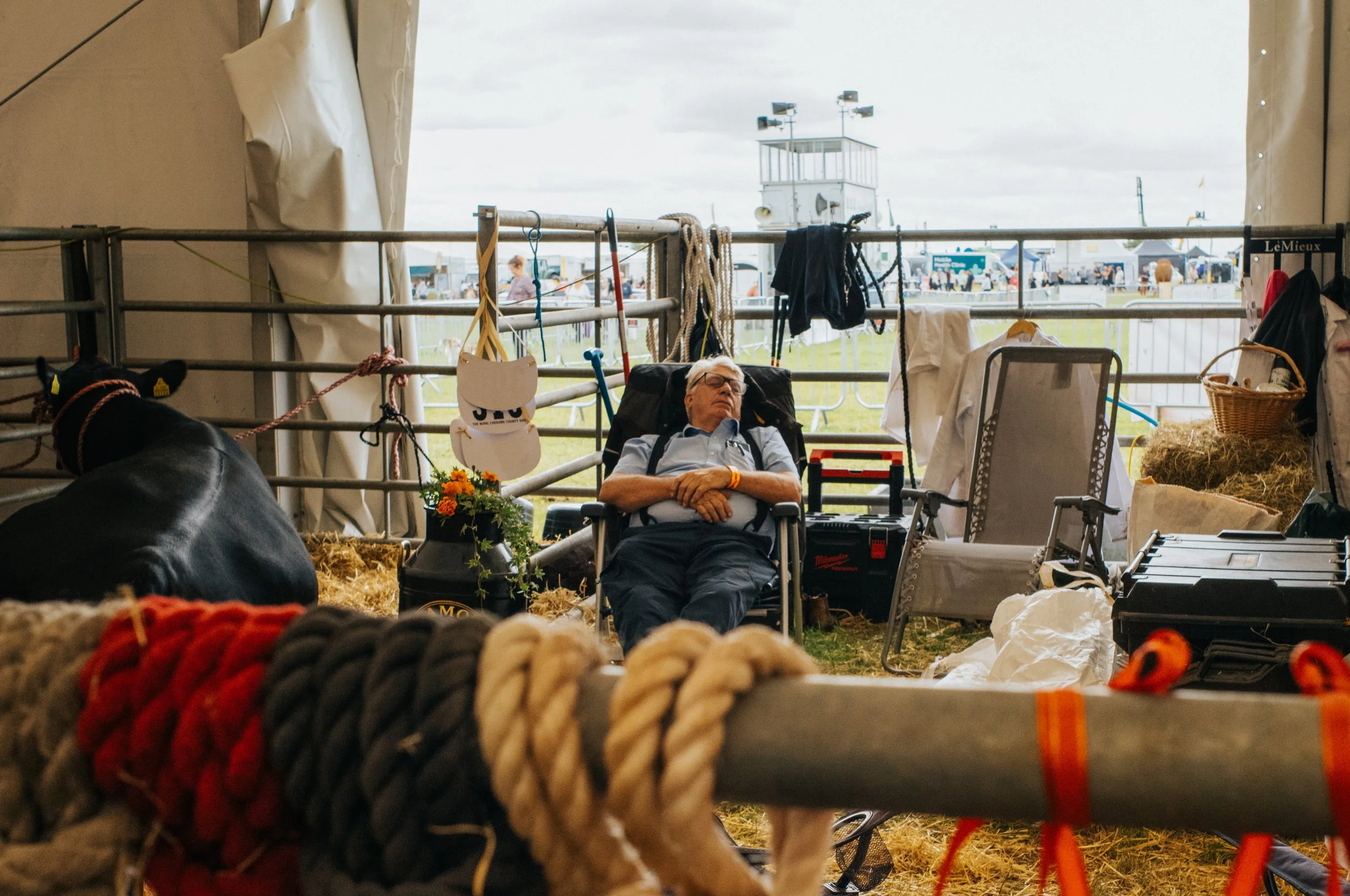 An elderly man with white hair and glasses naps in a folding chair inside a large tent, with various equipment and supplies nearby, at an outdoor event with tents and people in the background.