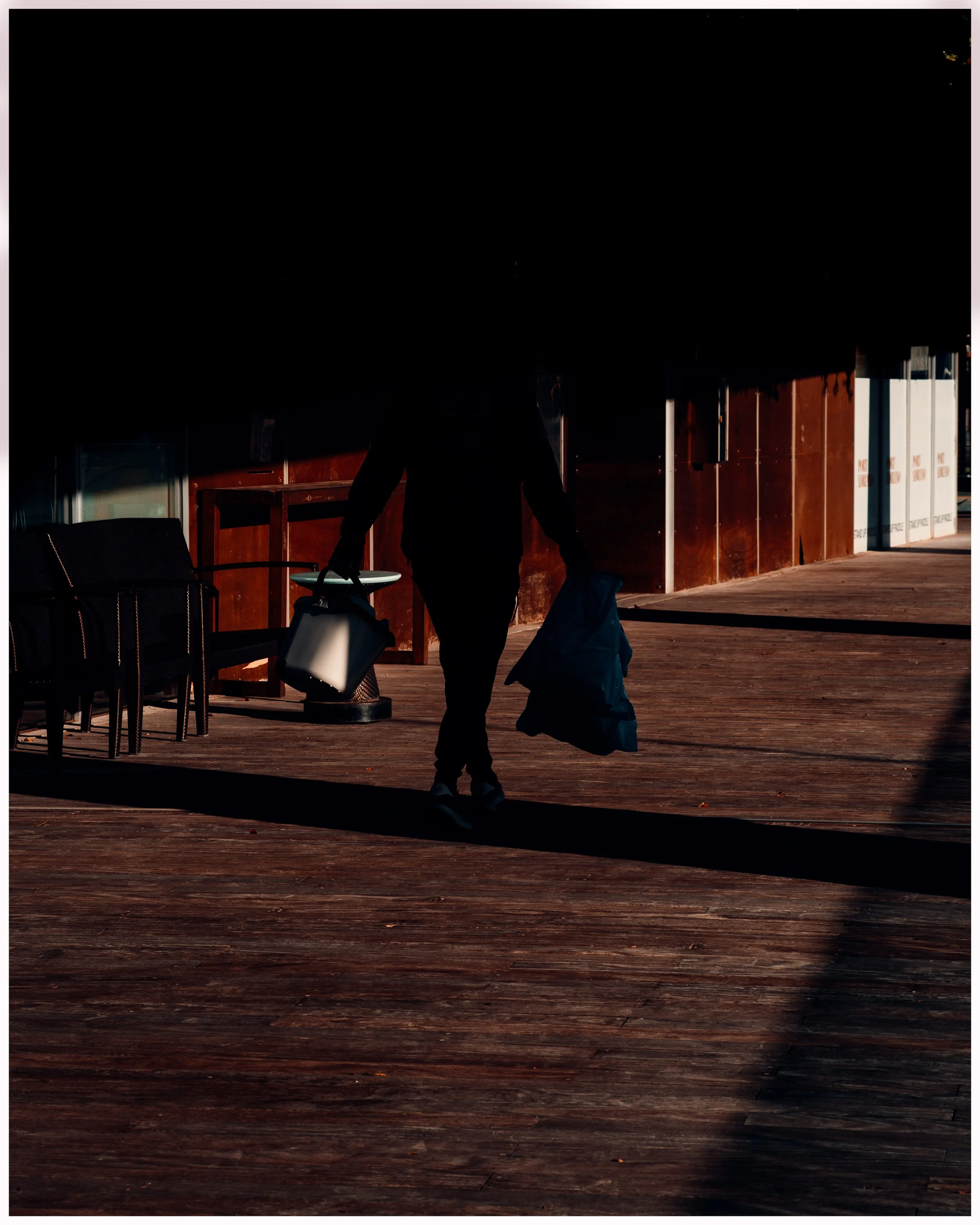 Silhouette of a person walking on a wooden deck carrying a shopping bag and a water jug, with sunlight casting long shadows.