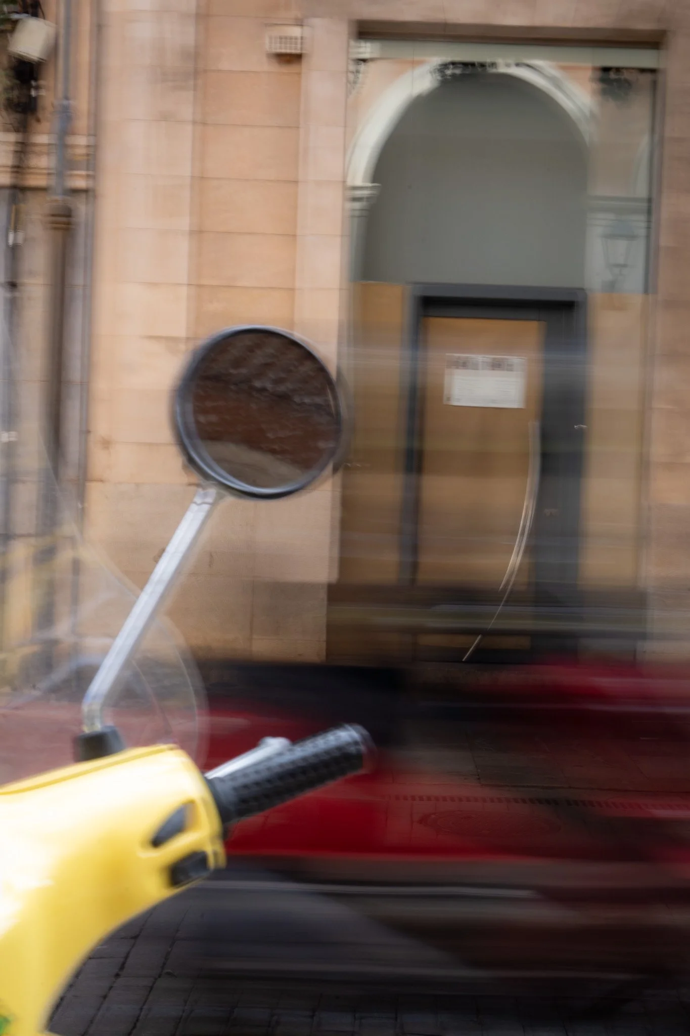A photo taken from a moving vehicle showing a yellow side-view mirror, part of a yellow vehicle, with a blurred building facade and a glass door in the background.