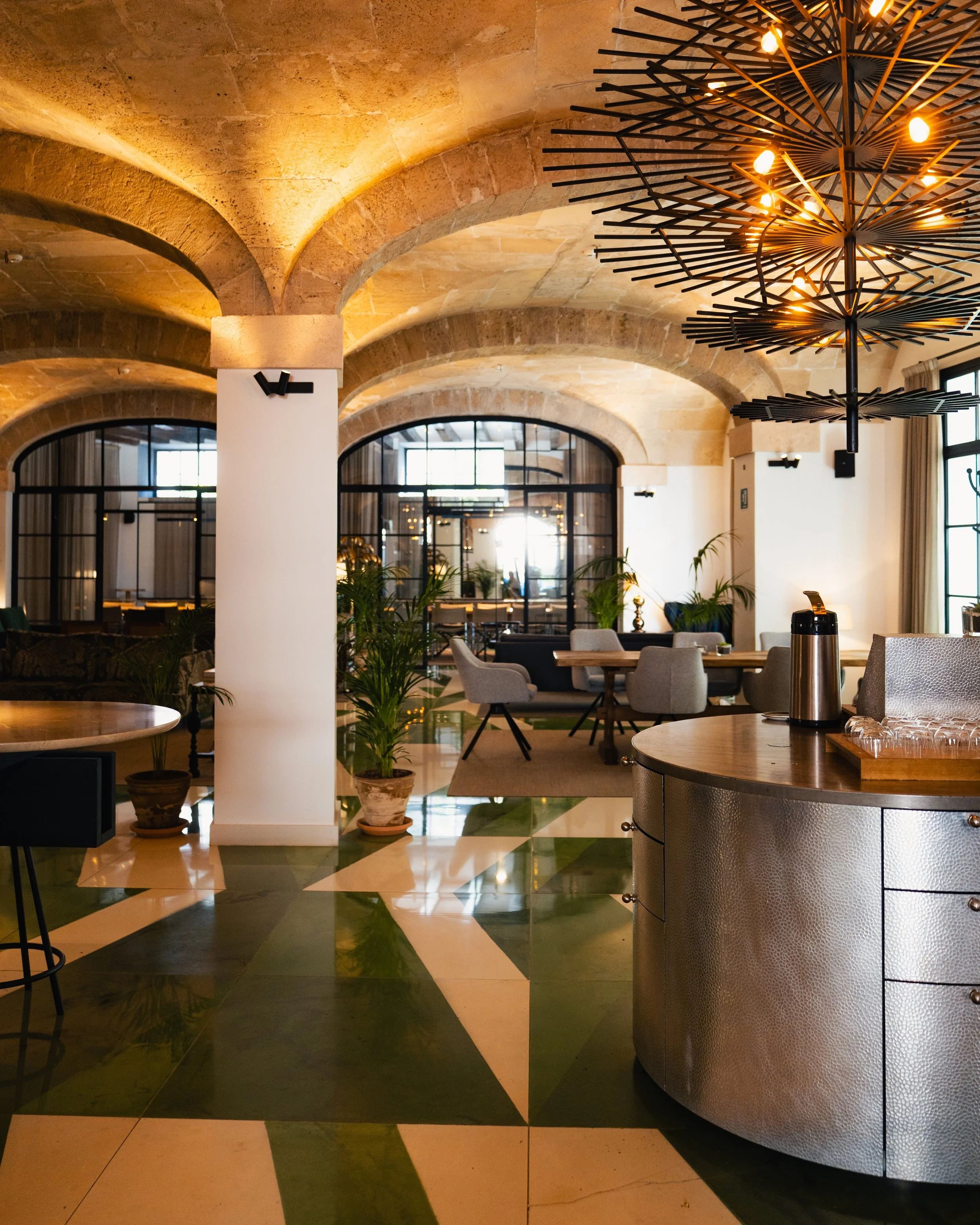 Elegant indoor restaurant or lounge with arched stone ceiling, modern chandelier, potted plants, and seating areas with tables and chairs.
