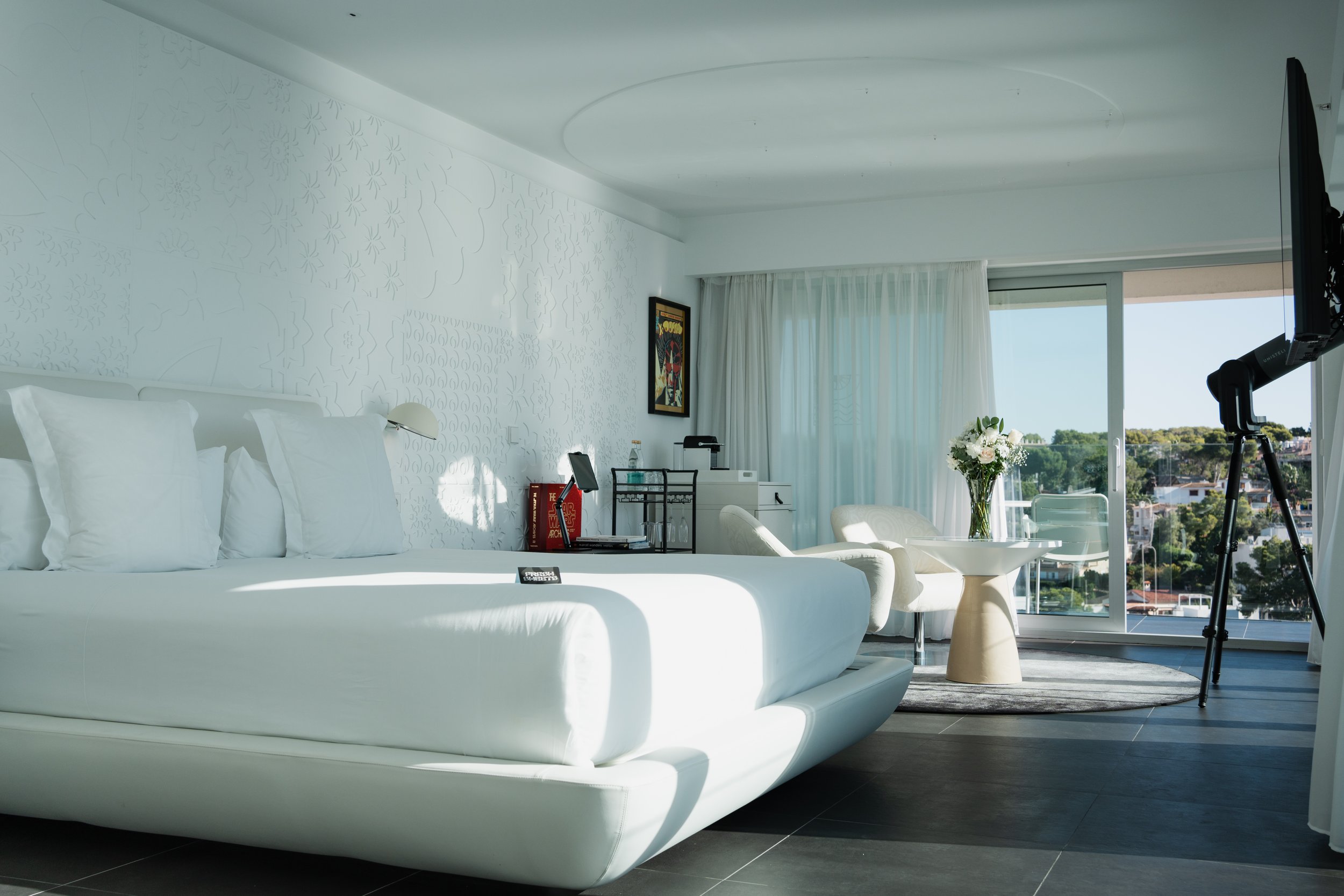 A modern, white hotel room with a large bed, table, chairs, a TV on a stand, and a sliding glass door leading to a balcony with a view of a cityscape and greenery, decorated with a vase of flowers and framed artwork.