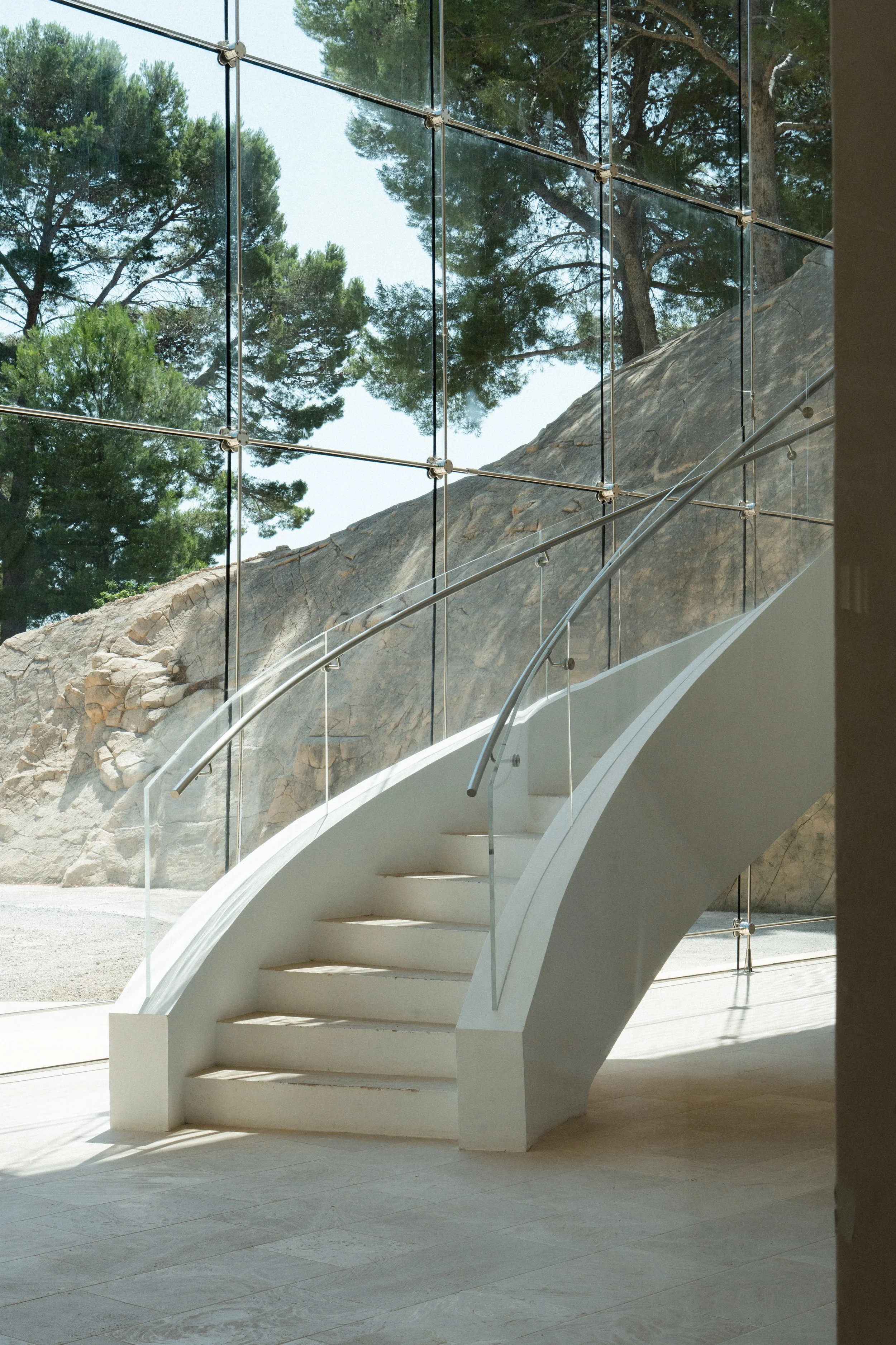 Modern white spiral staircase with glass and metal railing inside a building with large floor-to-ceiling windows looking outside at trees and rocky terrain.