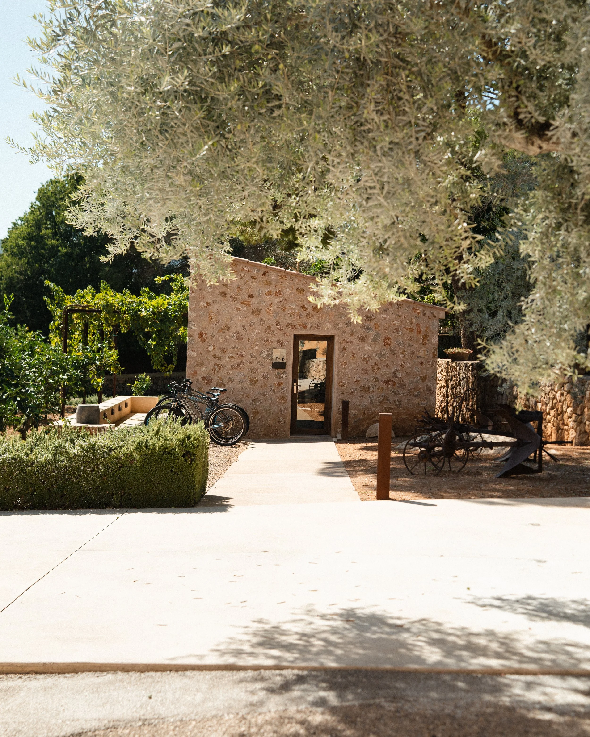Stone building with a glass door, bicycles parked to the left, and vintage farming equipment to the right, surrounded by trees and bushes.