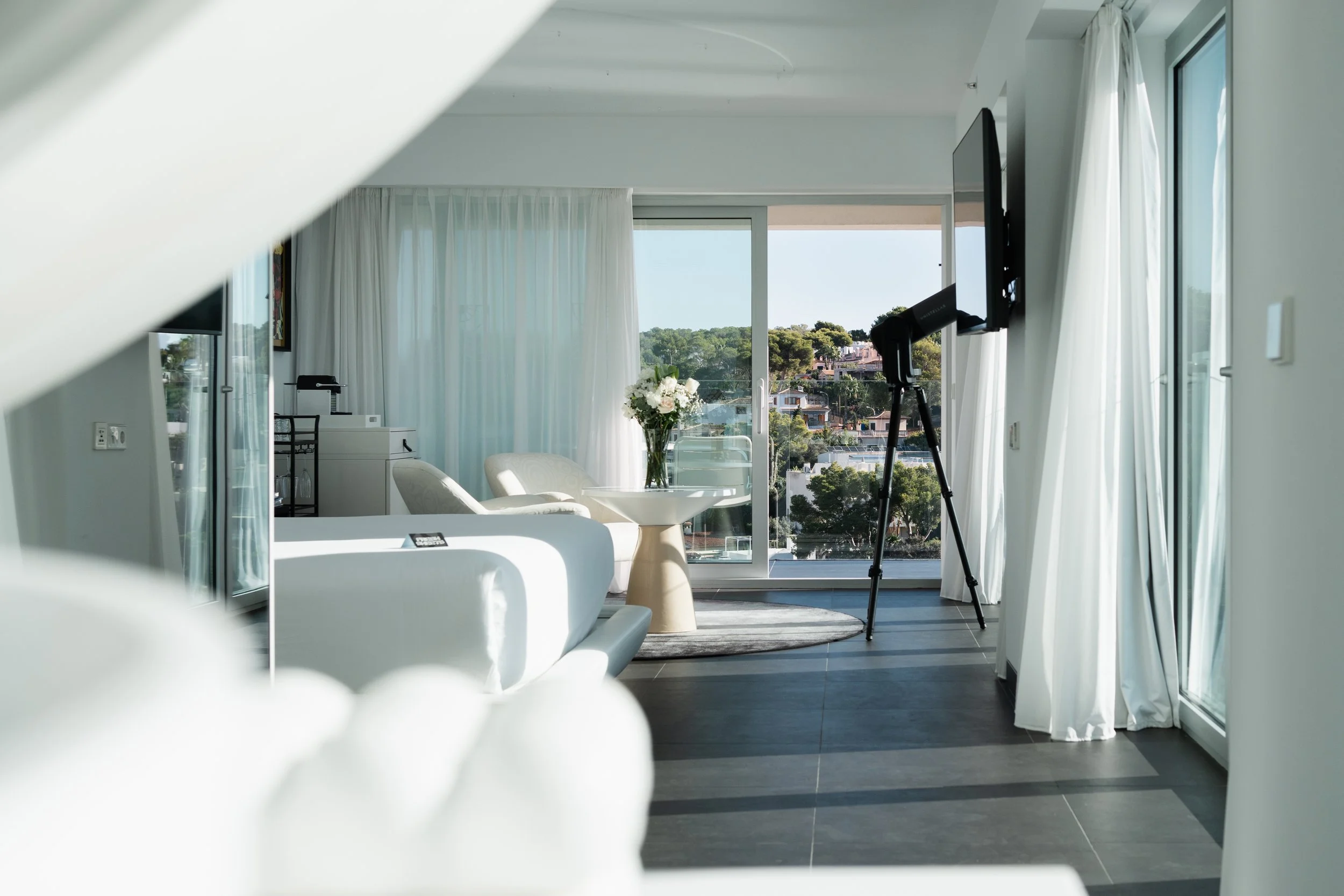 Modern living room with large windows, white curtains, white furniture, a vase with white flowers on a table, and a telescope facing the window.