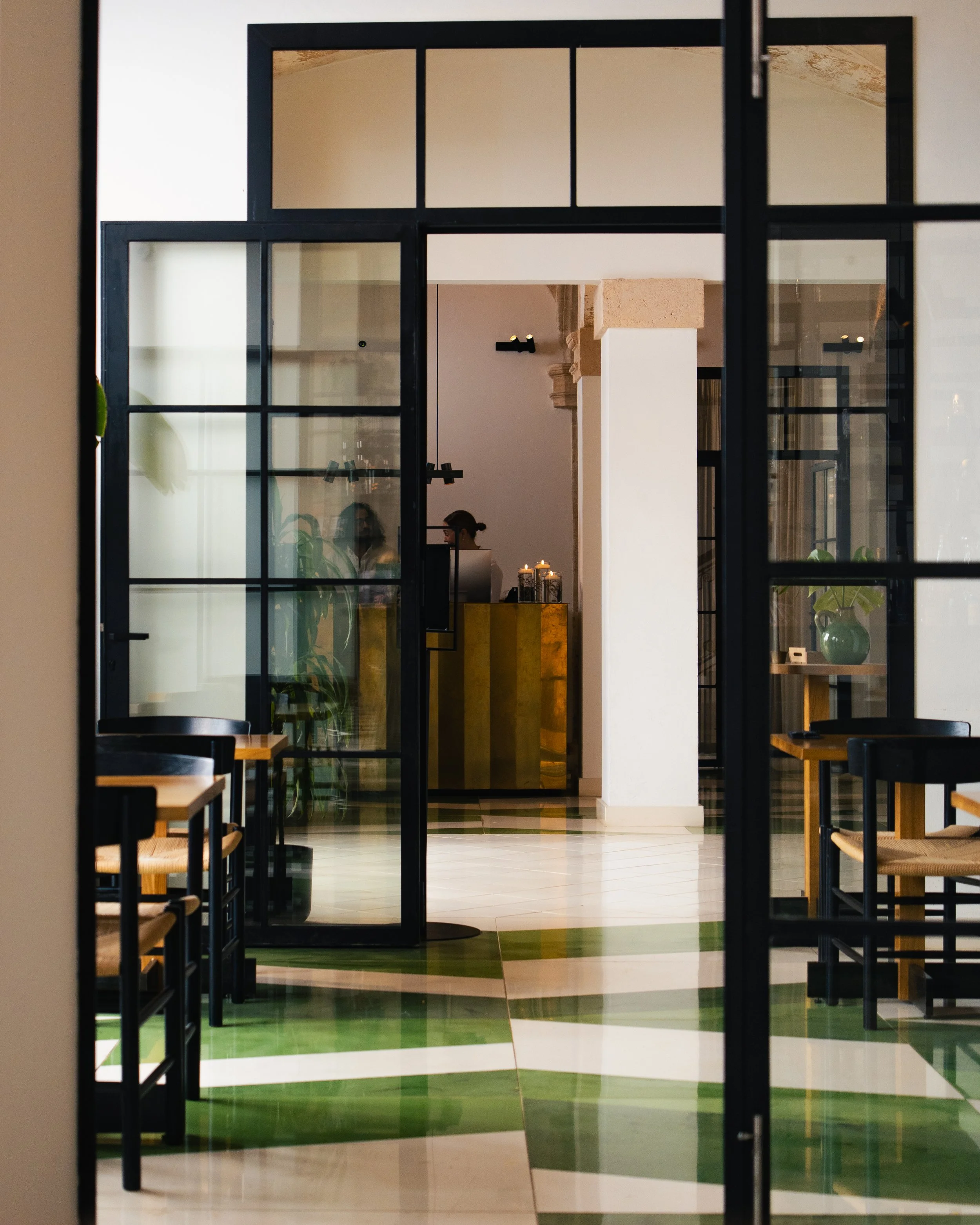 View through glass partitions into a stylish cafe with a reception counter, seating area, and candles on the counter.