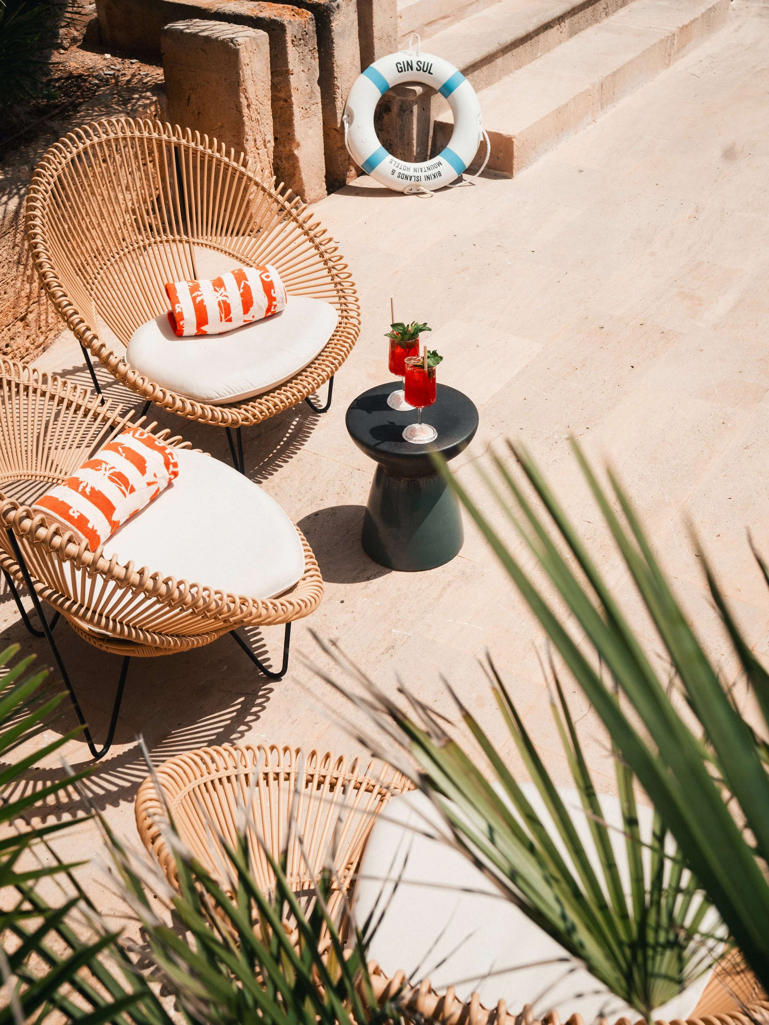 Outdoor patio seating with two rattan chairs with white cushions and orange-striped towels, a black side table with two red drinks garnished with mint, and a life preserver with the words 'GIN SUL' on it, set against a stone wall and stairs.