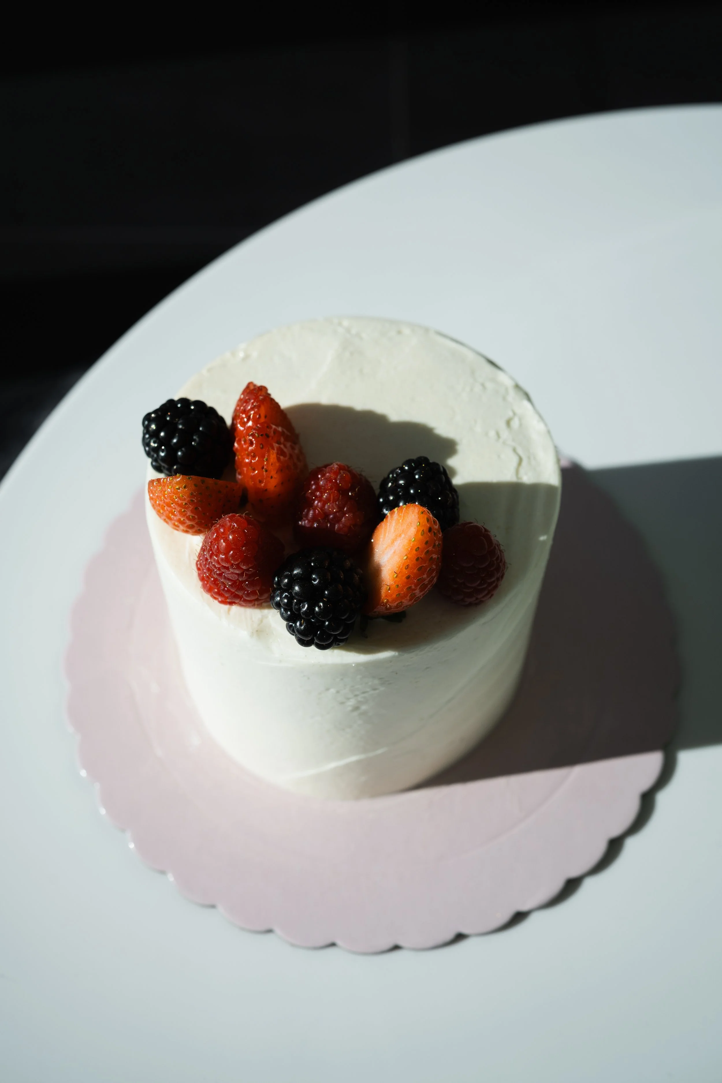 Close-up of a round cheesecake topped with mixed berries including blackberries, strawberries, and raspberries, placed on a white scalloped-edged plate.