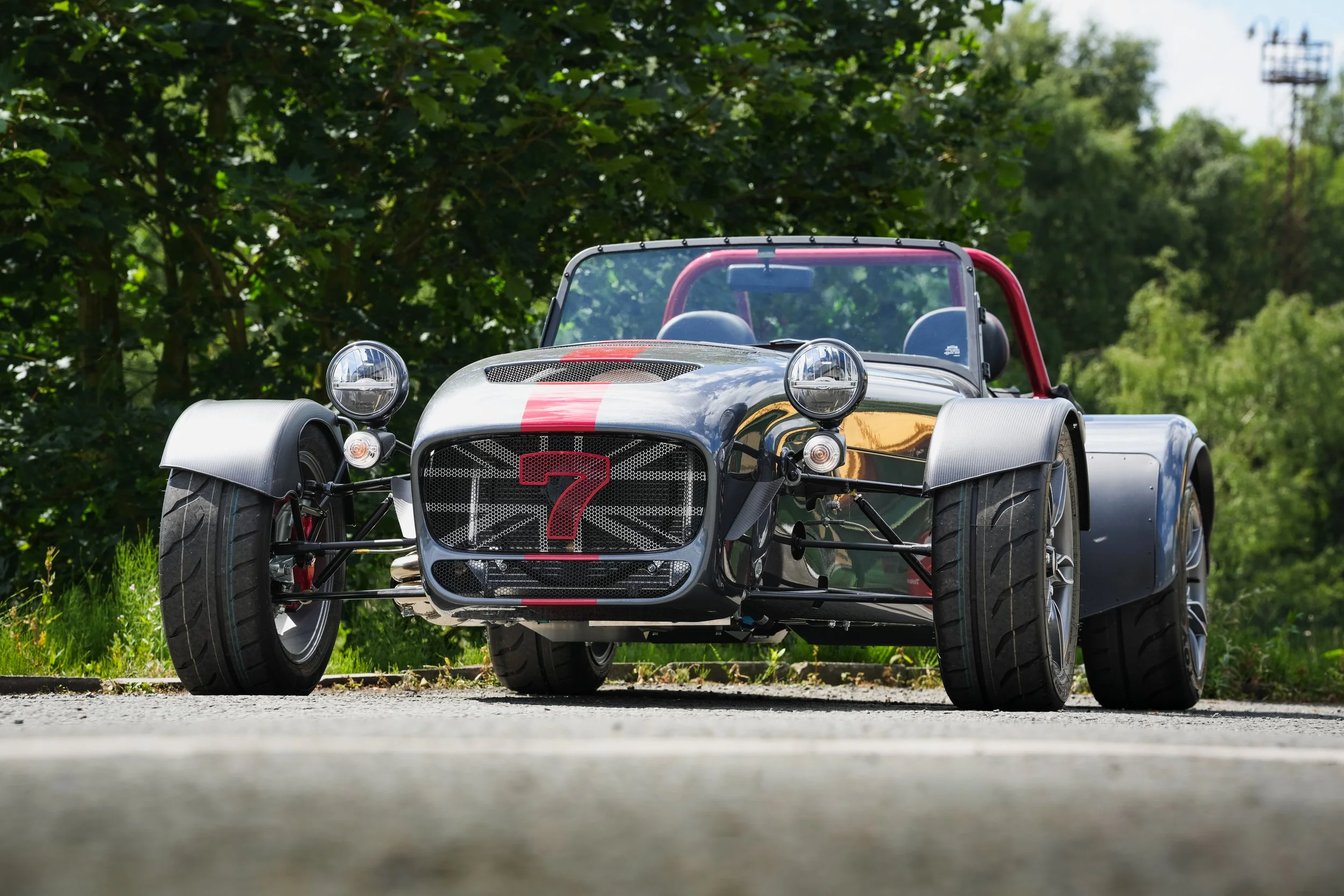 A silver open-wheel race car with a red stripe and a large red number seven on the front, parked outdoors on a grassy area with trees and blue sky in the background.