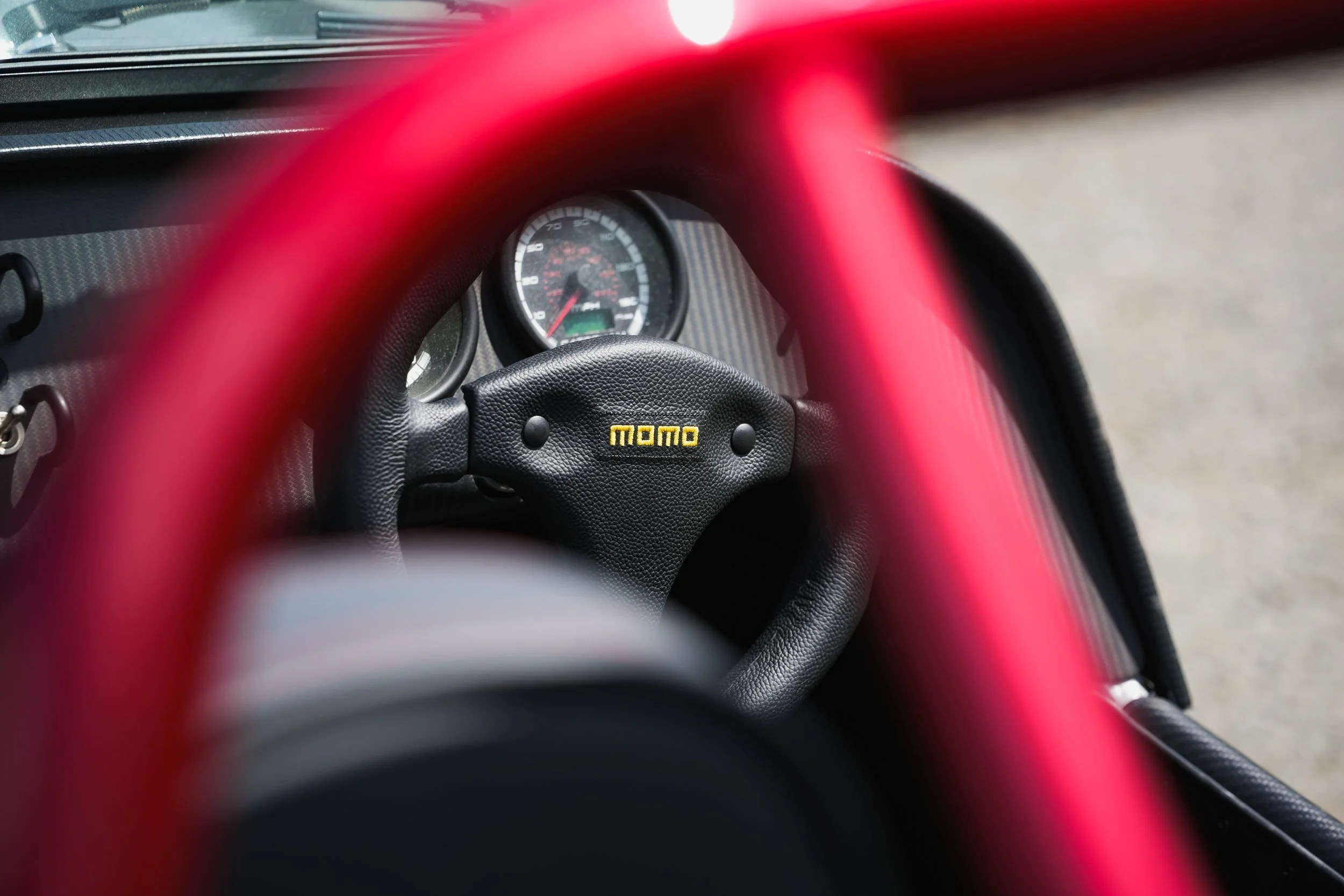 Close-up view of a race car dashboard with a Momo steering wheel and red roll cage, showing speedometer and tachometer gauges, with blurred red racing harness in the foreground.