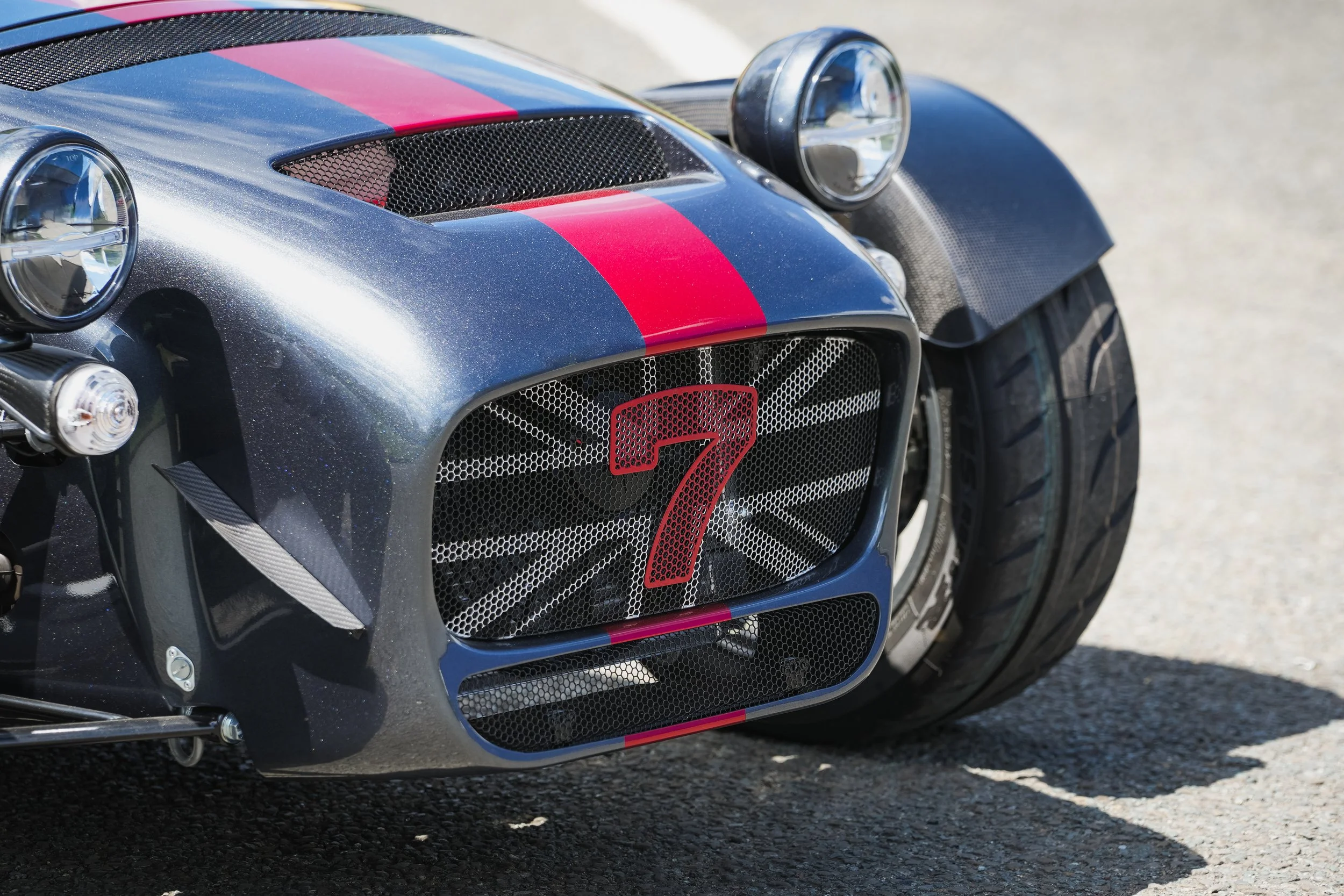 Close-up of a race car's front end with a metallic gray body, pink racing stripe, round headlamps, and a black mesh grille with red number 7