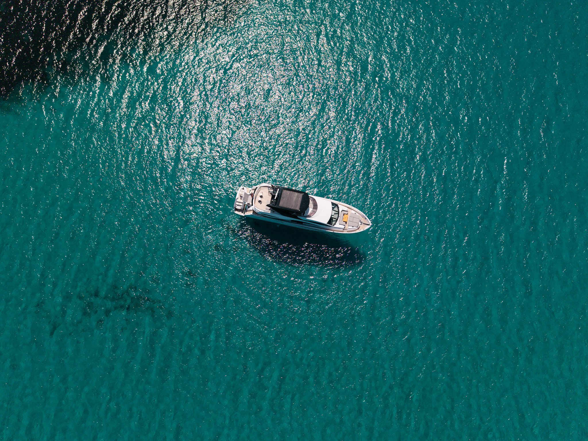 An aerial view of a boat floating on calm, teal water, casting a shadow.