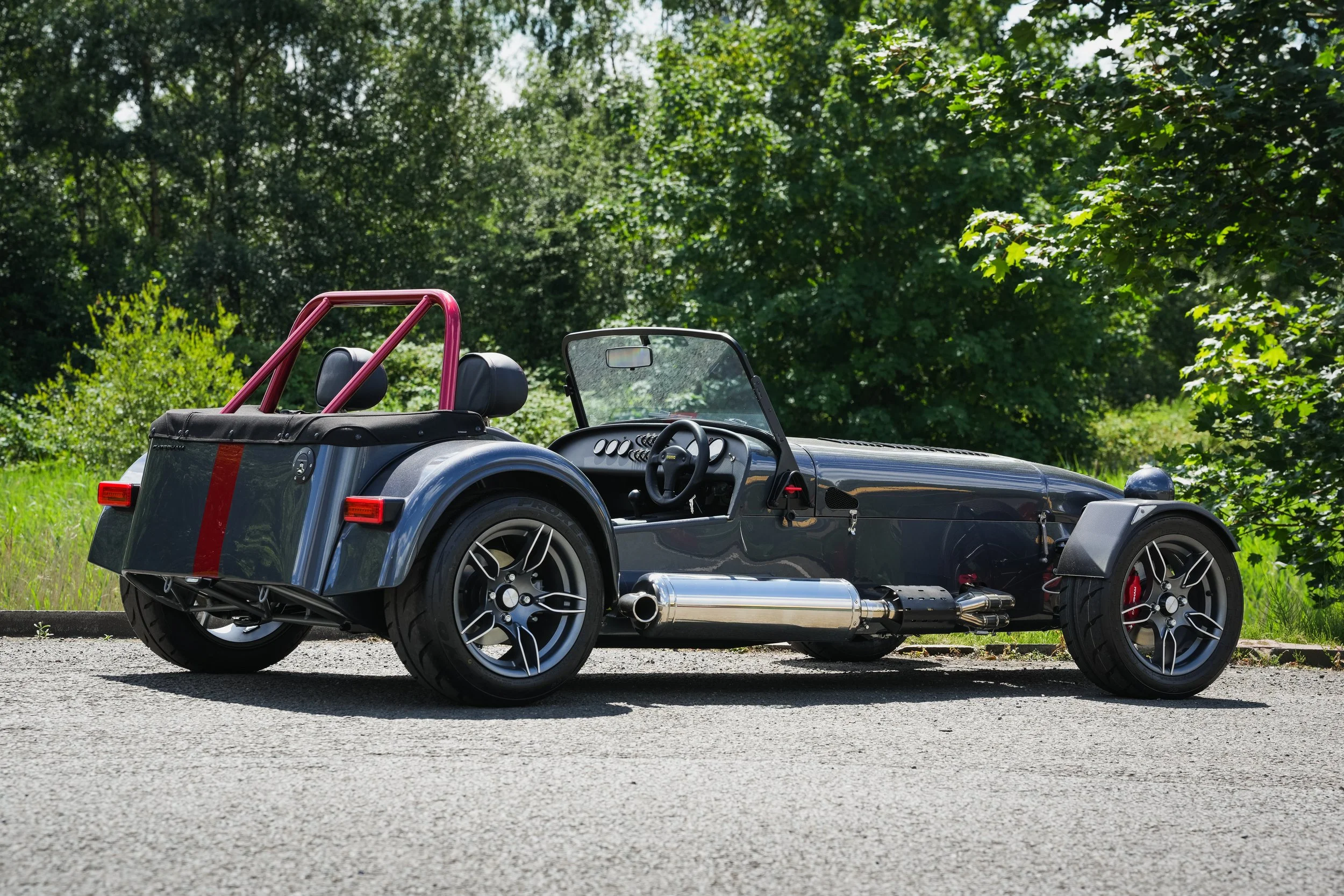 A vintage-style sports car parked on a roadside with lush green trees in the background.