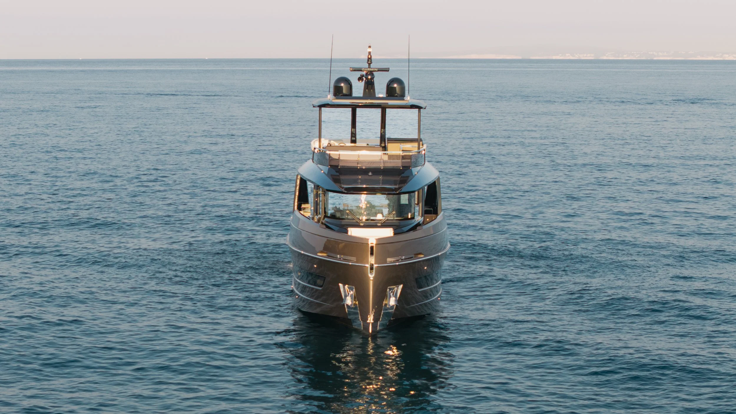 A luxury yacht sailing toward the camera on calm ocean waters with a distant shoreline in the background.