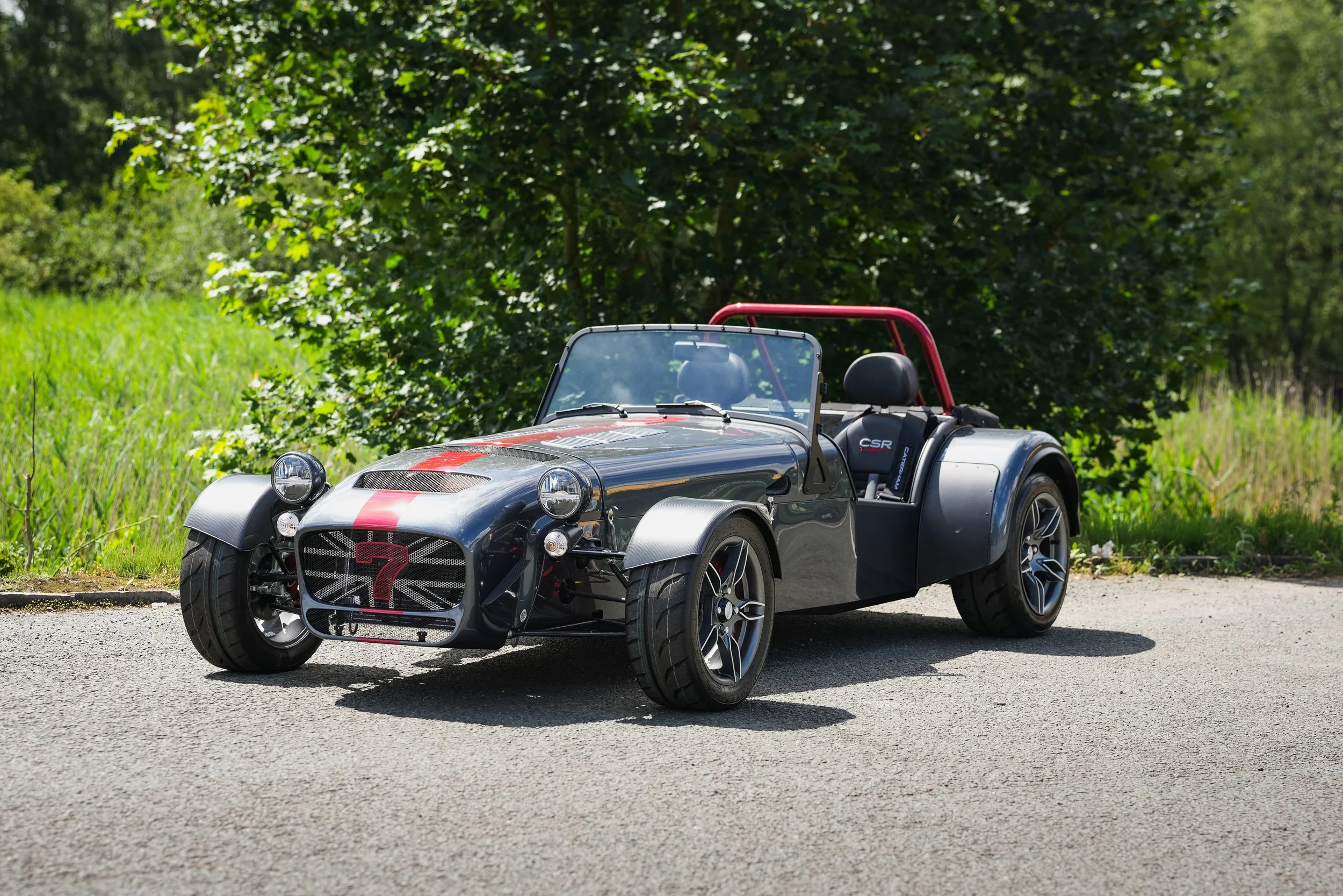A small black and gray sporty open-top car with a red racing stripe, parked on a paved surface amidst green grass and trees.