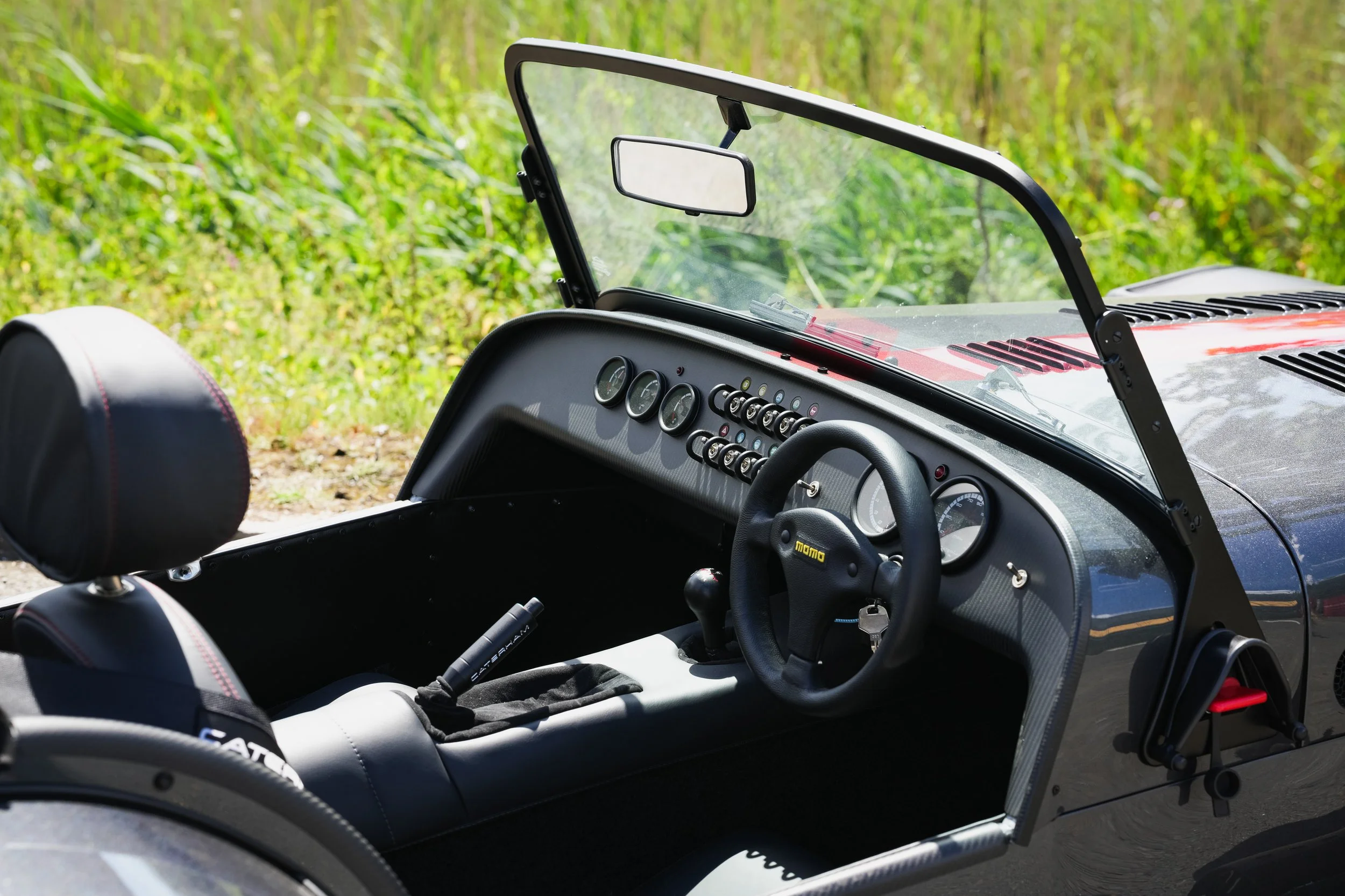 Interior of a vintage race car with a dashboard, steering wheel, and other controls, parked on a dirt path next to tall green grass.