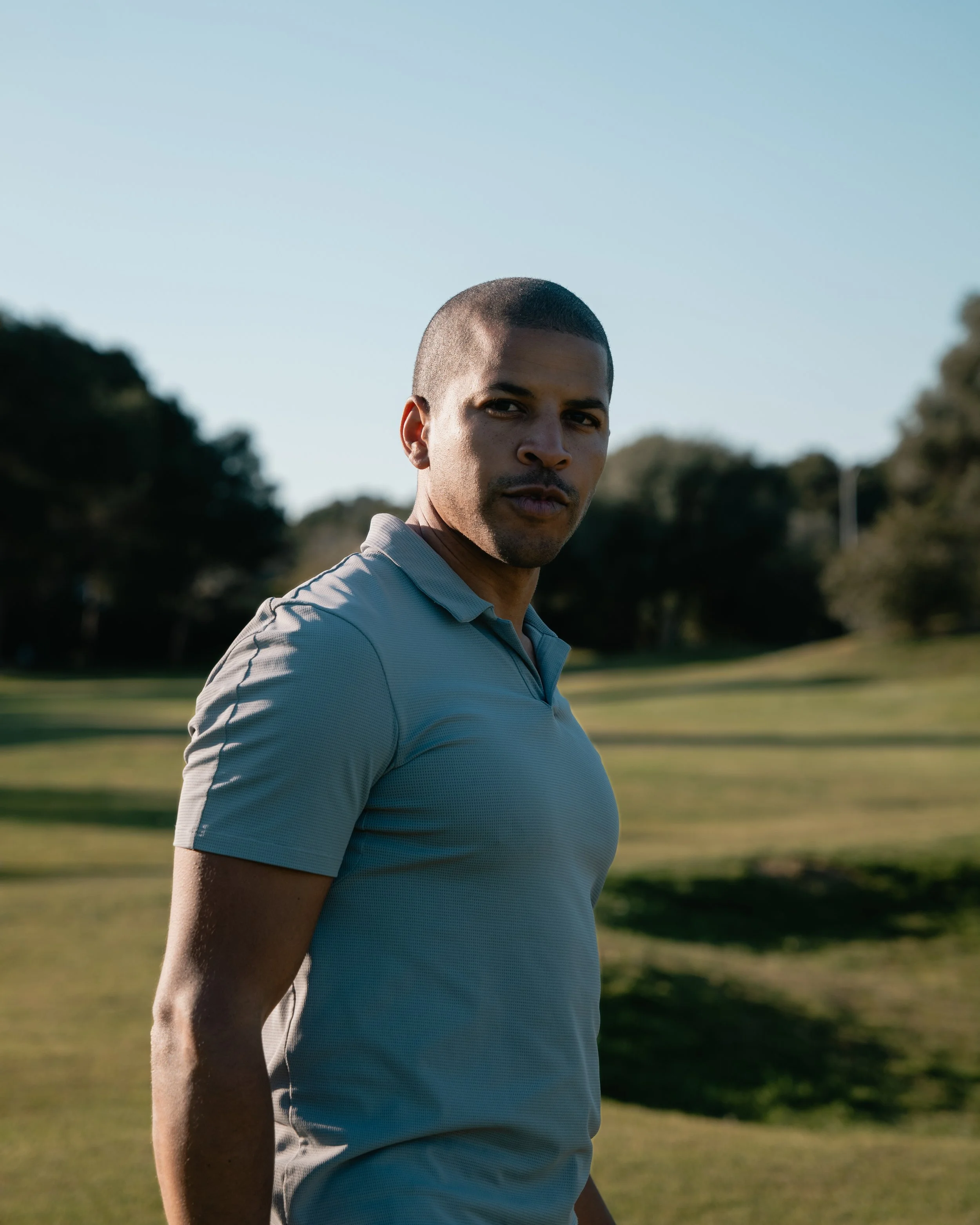 A man standing outdoors on a golf course, wearing a light gray polo shirt, with trees and a clear blue sky in the background.