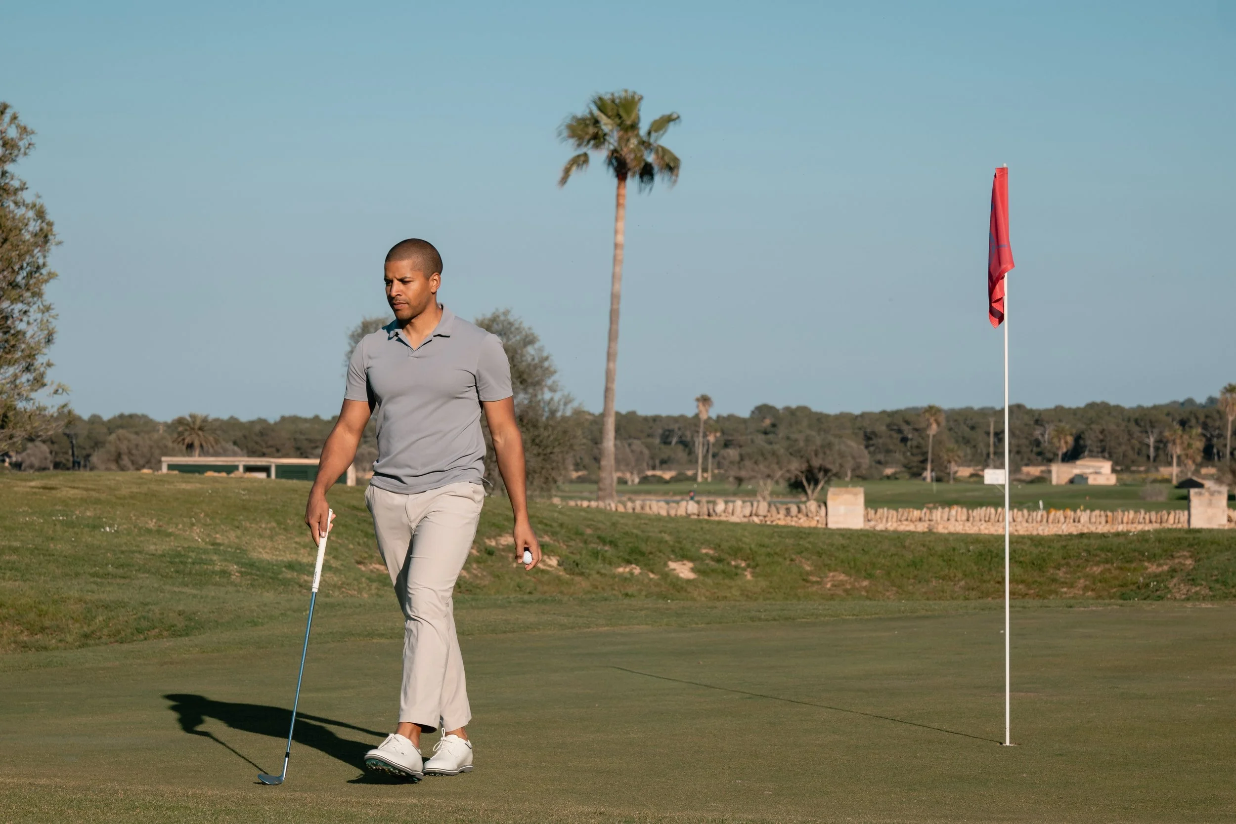 A man walking on a golf course holding a golf club in his right hand near the putting green, with a red flag in the hole behind him, under a clear blue sky, with palm trees and bushes in the background.