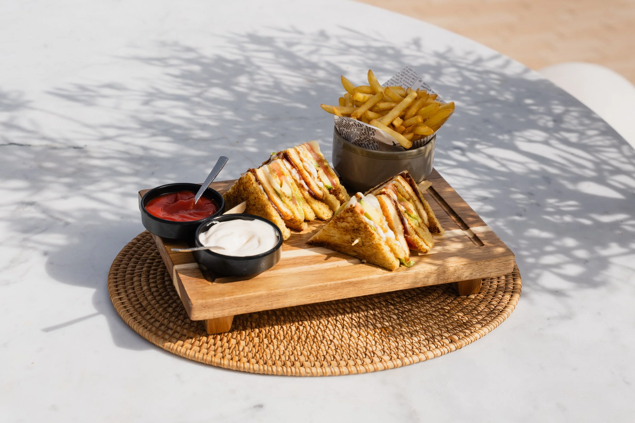 A wooden tray holds a sandwich cut into triangular halves, with visible layers of lettuce, tomato, and cheese, accompanied by small bowls of ketchup, mayonnaise, and a white sauce. Next to the sandwich are a metal cup of French fries in newspaper-lin