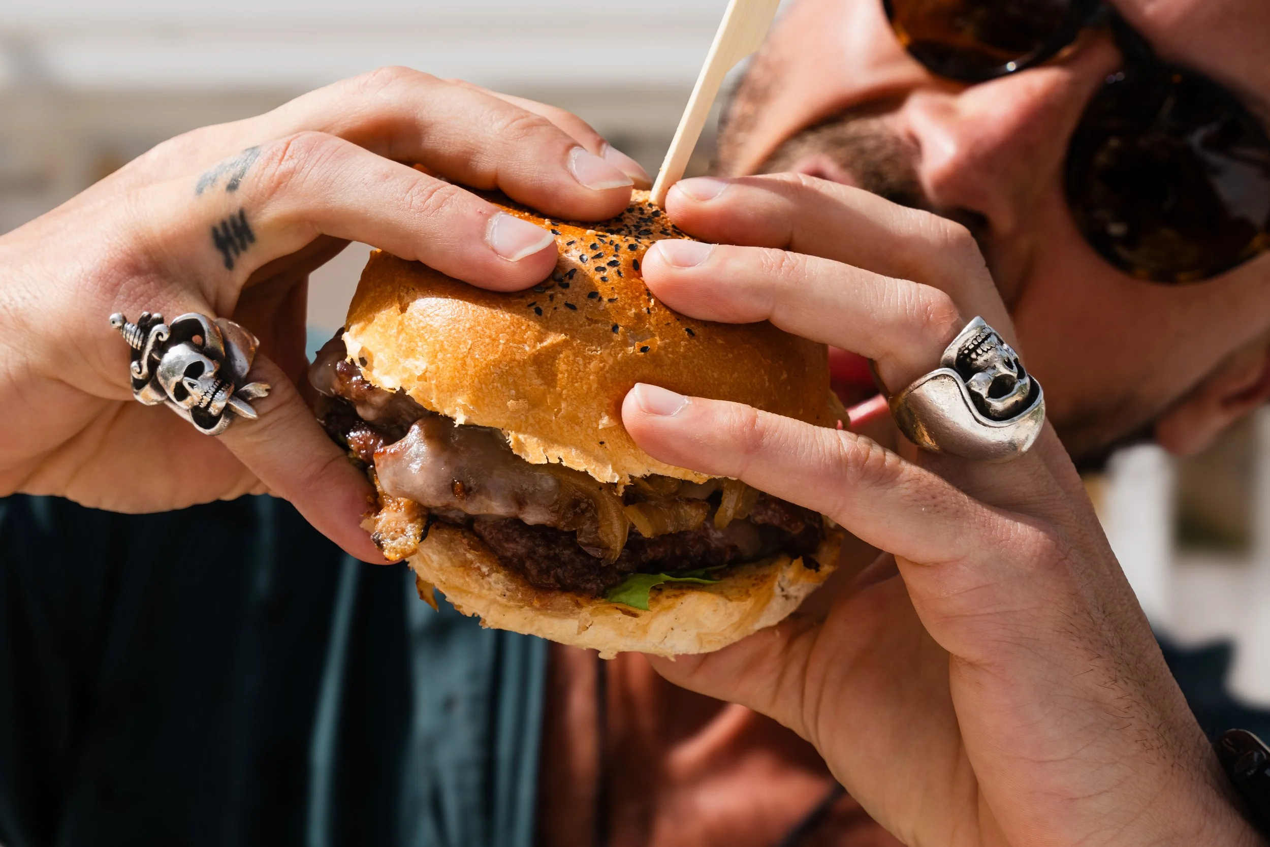 Person wearing sunglasses and silver jewelry bites into a cheeseburger with black sesame seeds on the bun.