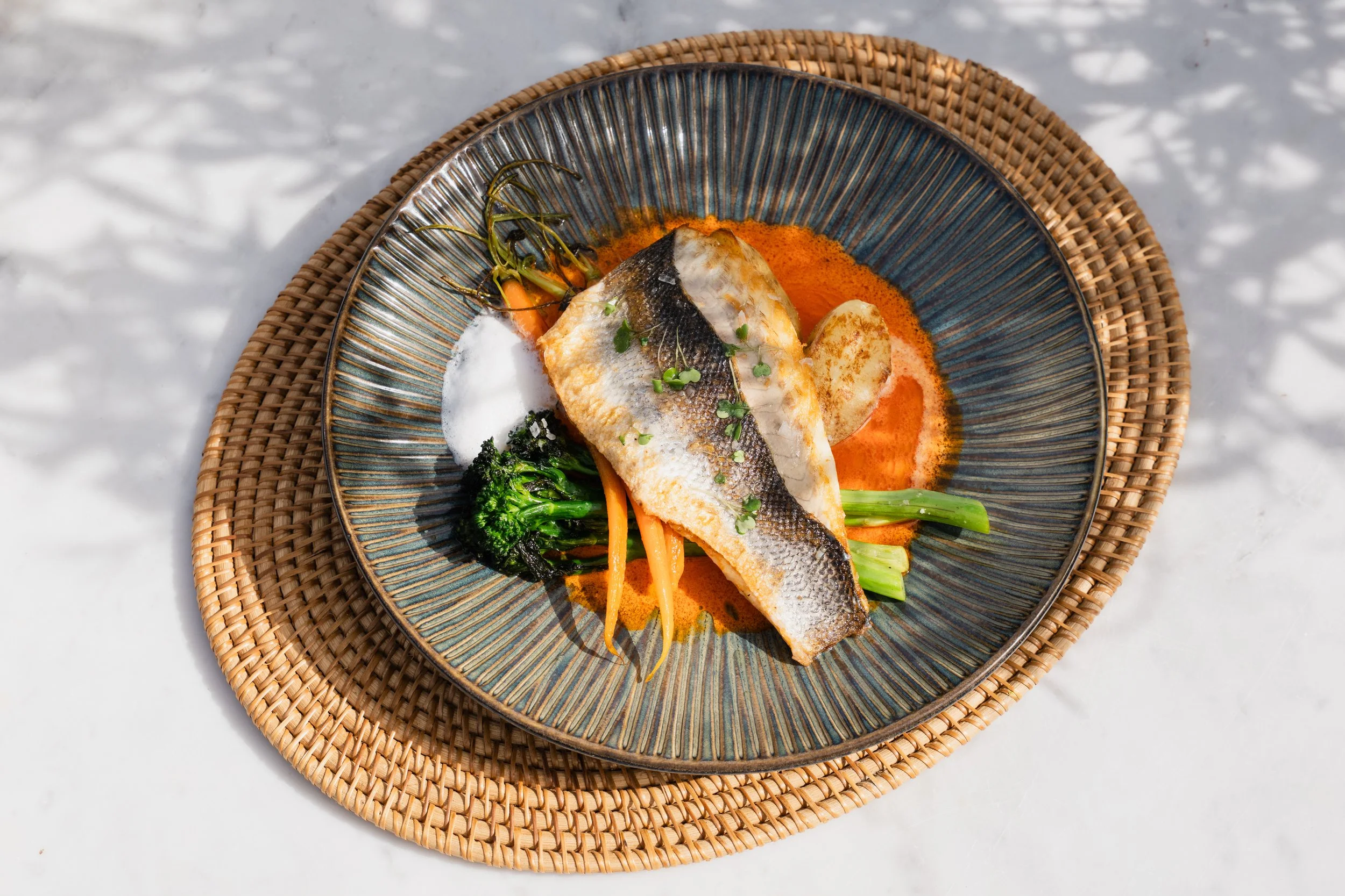 A plate of cooked fish fillet with sauce, green vegetables, and carrots on a decorative bowl placed on a woven placemat on a white surface outdoors.