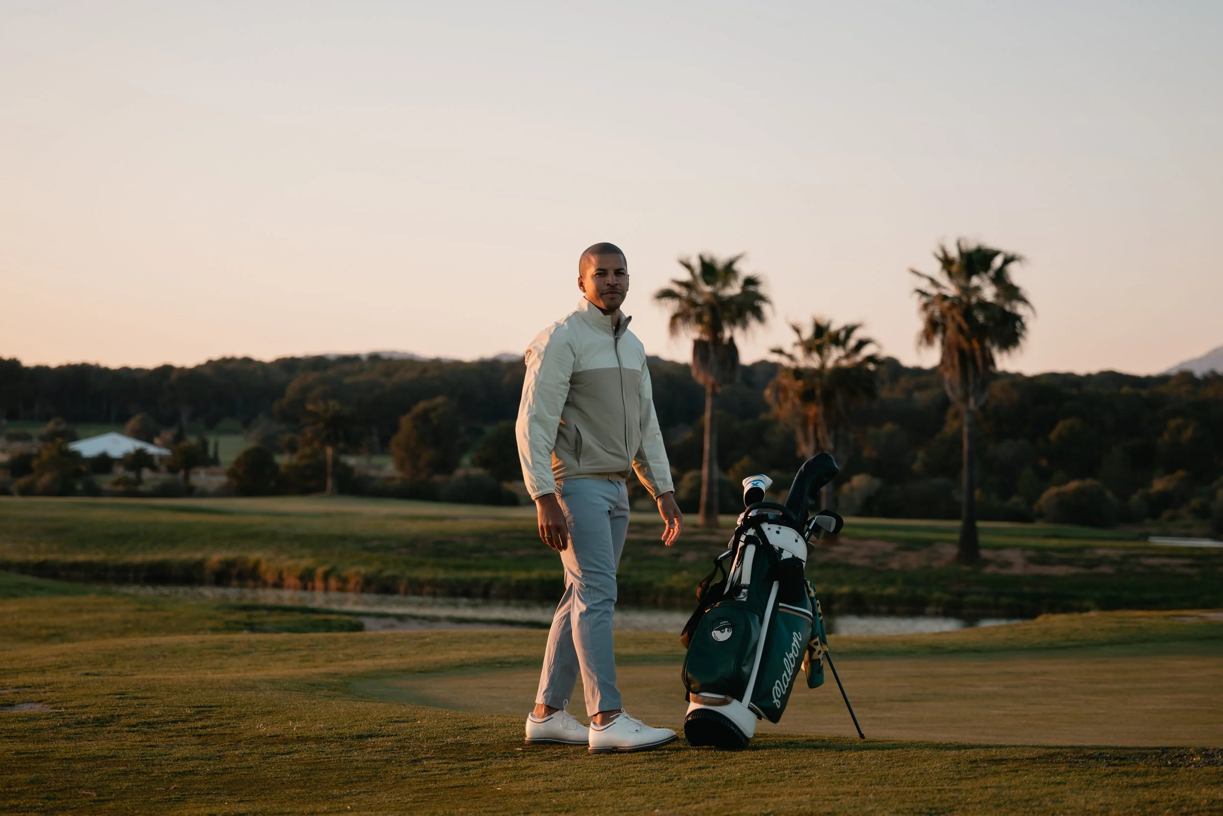 A man standing on a golf course holding a golf club next to a golf bag during sunset with palm trees and hills in the background.
