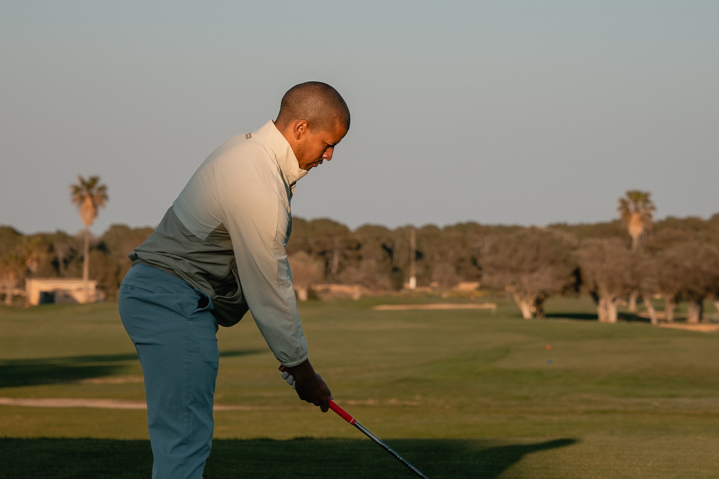 A man wearing a light-colored jacket and blue pants is preparing to hit a golf ball on a golf course during sunset. He is holding a golf club and looking down at the ball with a focused posture, surrounded by a landscape with trees and a clear sky.