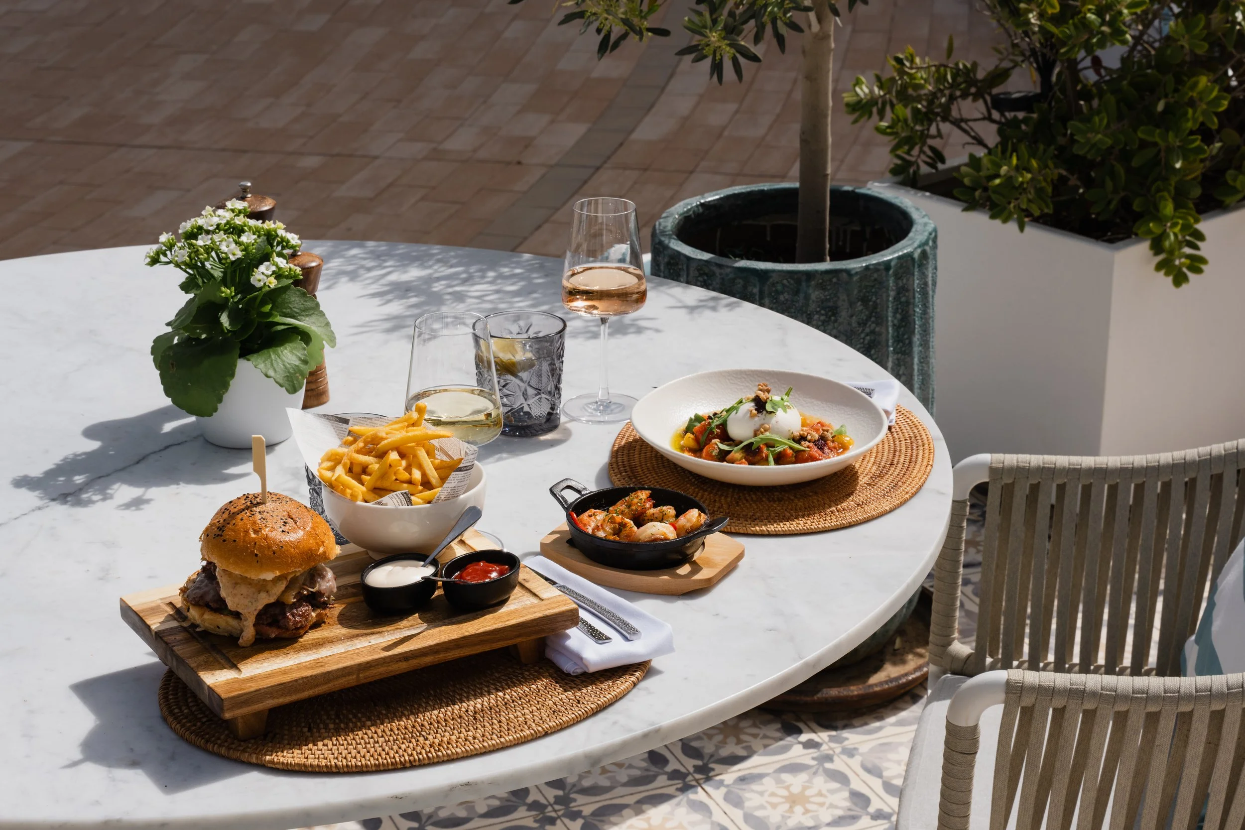 An outdoor dining table set with various foods and drinks, including a burger on a wooden board, a bowl of fries, a glass of white wine, a glass of rose wine, a dish with vegetables and a poached egg, and a small cast iron skillet with shrimp, on a w