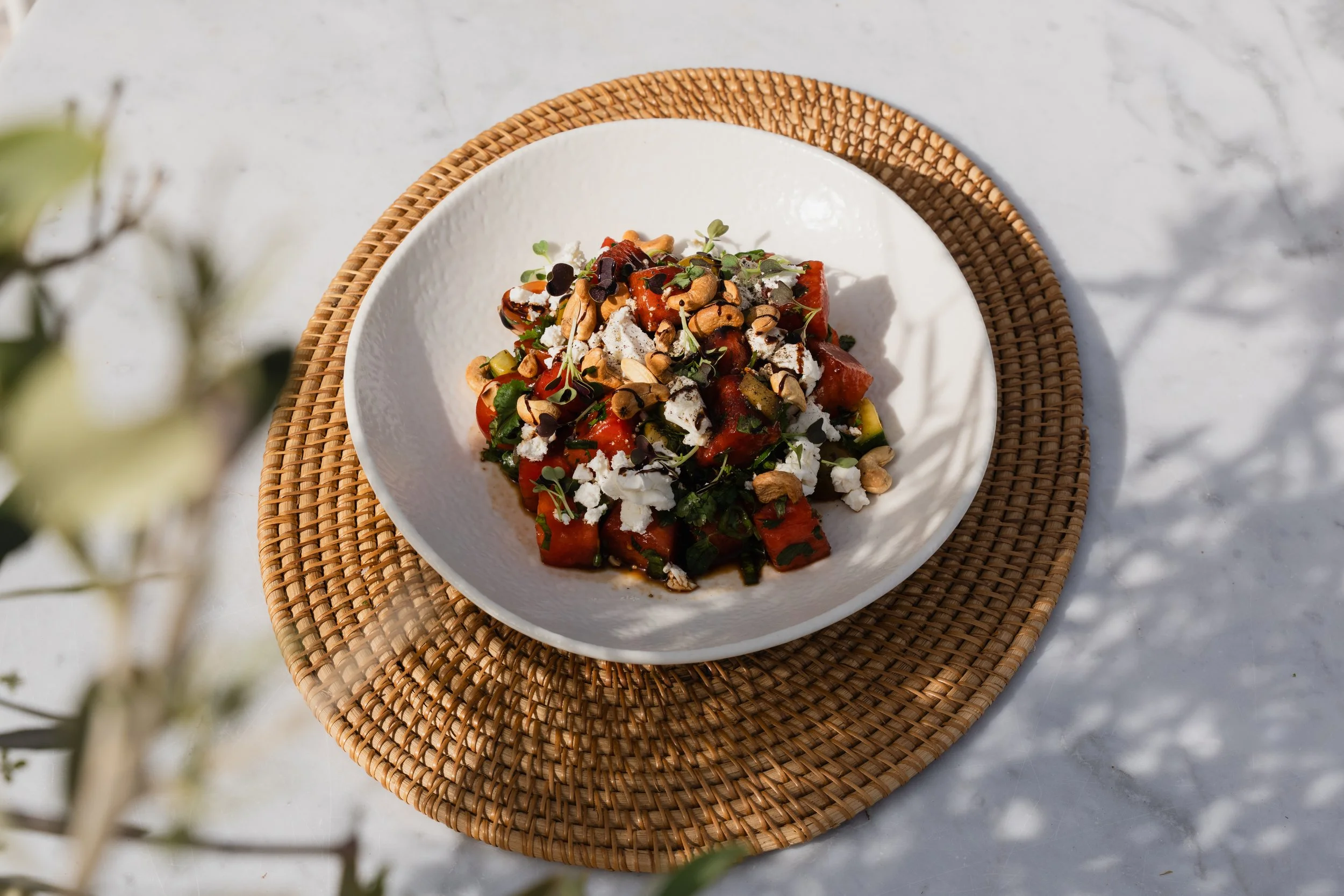 A white bowl with a textured surface containing a salad with watermelon, feta cheese, nuts, herbs, and a drizzle of balsamic glaze, placed on a woven placemat on a marble surface.