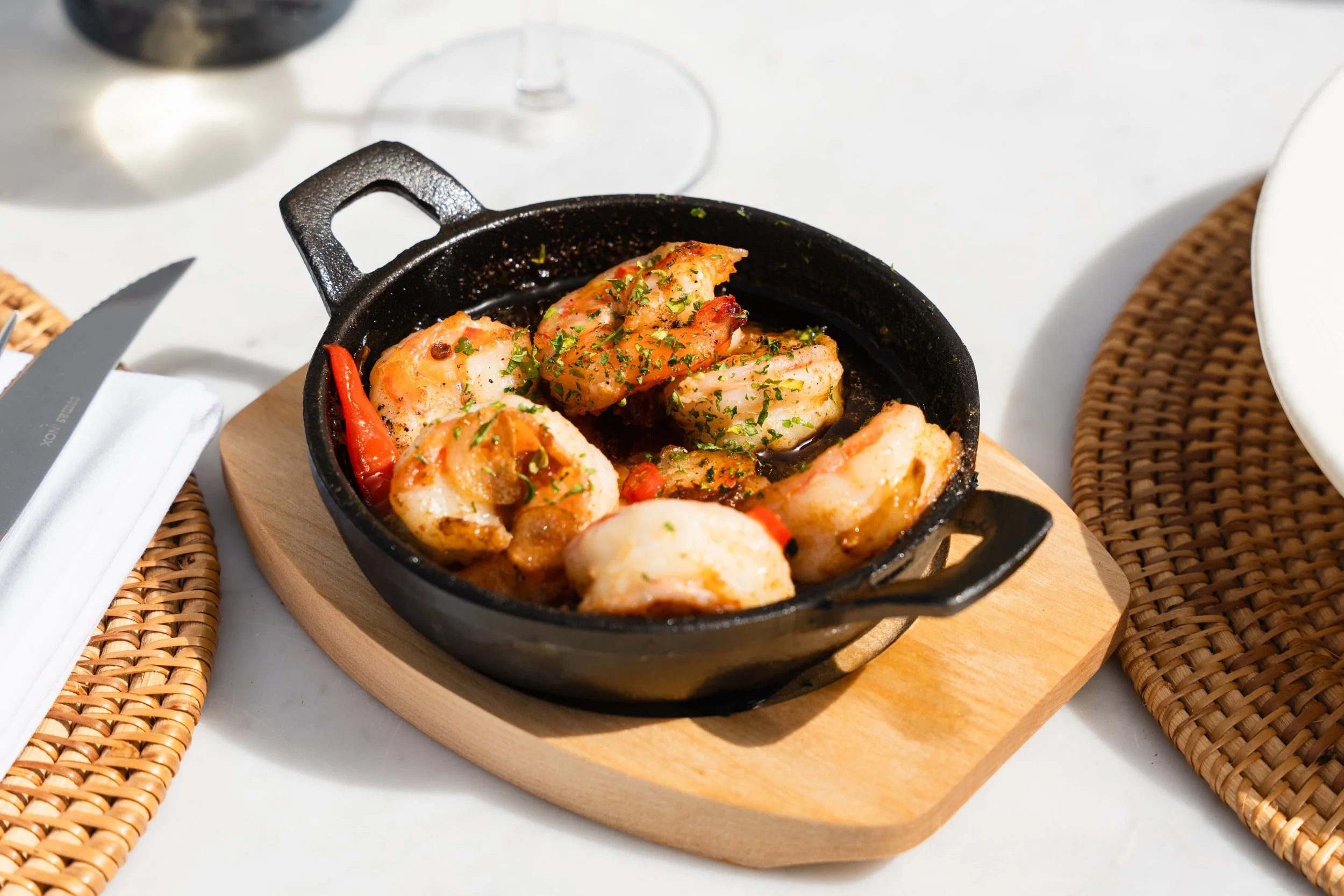 Cooked shrimp garnished with herbs in a small cast iron skillet on a wooden serving board, with a glass of red wine and table setting nearby.