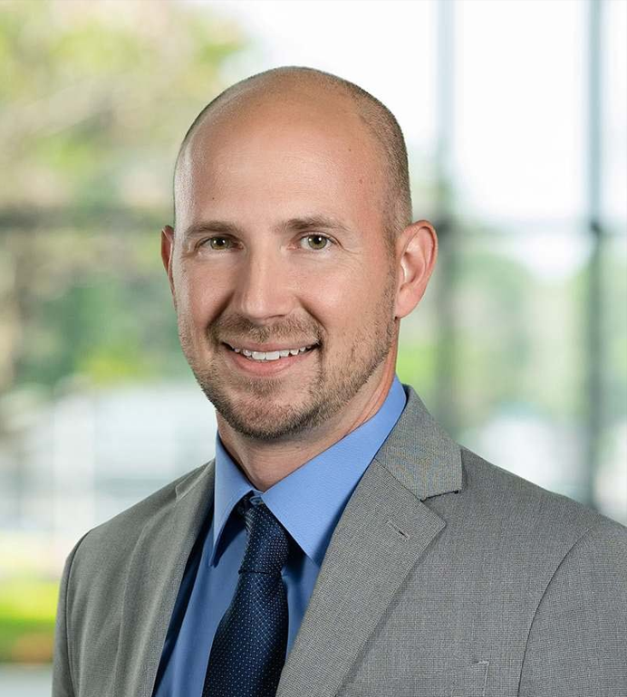 Professional headshot of a smiling man wearing a gray suit, blue shirt, and tie, with a blurred window background.