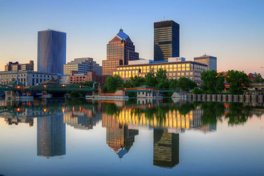 City skyline with tall buildings reflected in a calm river at sunset, including a mix of modern and historic architecture, trees, and a bridge.