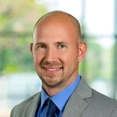 Professional headshot of a smiling man wearing a gray suit, blue shirt, and tie, with a blurred window background.