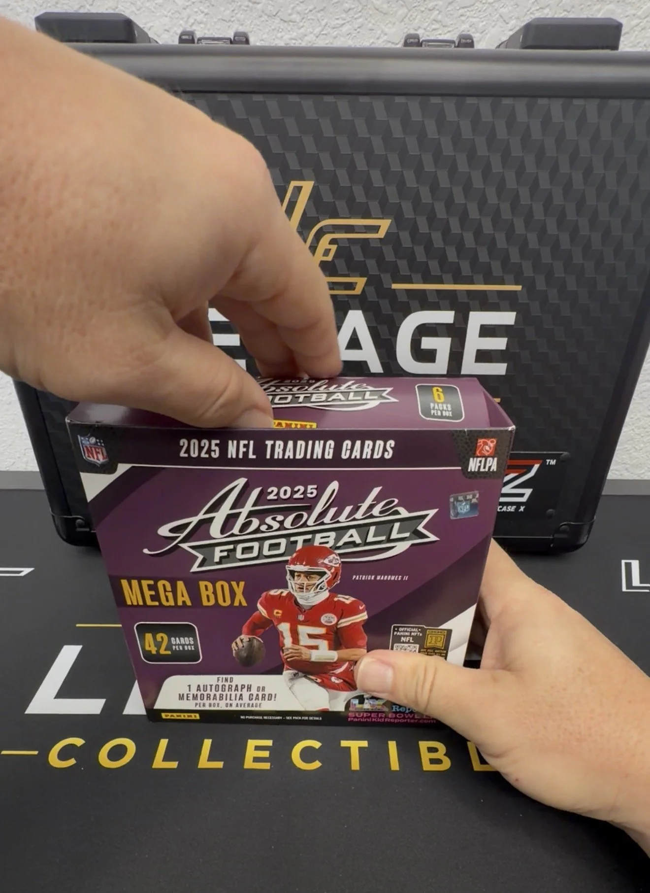 A person holding a box of 2025 NFL trading cards called Absolute Football Mega Box with an image of Patrick Mahomes on the front. In the background, a hand is inserting the box into a black case labeled "Collectible".