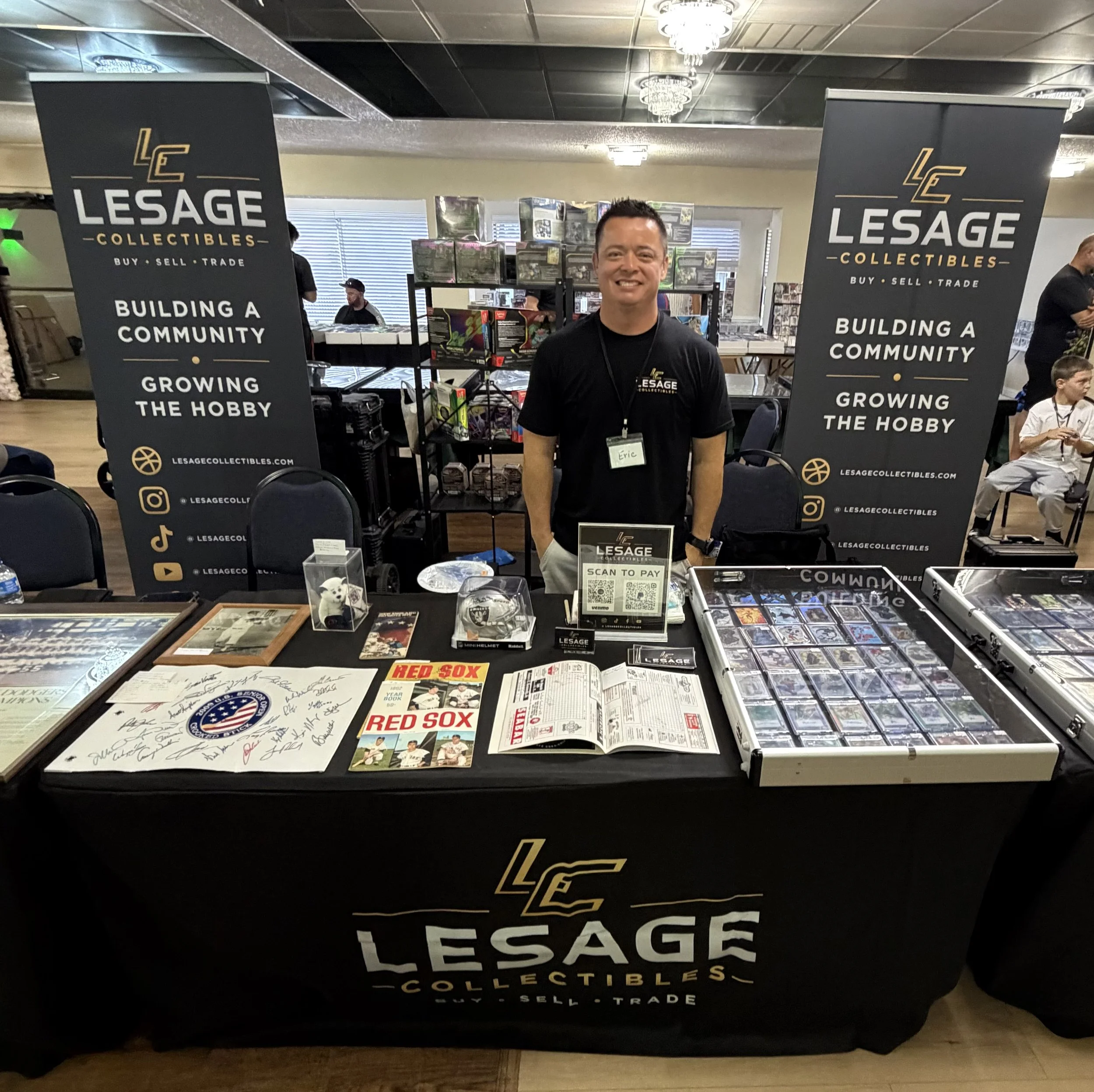 At an event booth for Lesage Collectibles, a man wearing a black shirt with a name tag standing behind a table displaying collectibles, with banners on either side that say 'Lesage Collectibles' and promote buying, selling, and trading collectibles to build a community and grow the hobby. The table has various items, documents, and display cases with collectibles, and there are other people in the background.