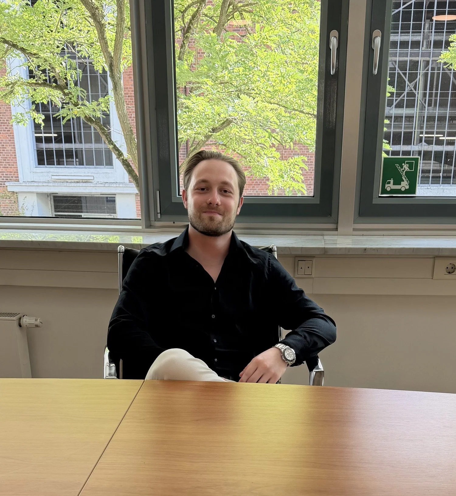 Ein junger Mann sitzt entspannt an einem Tisch im Büro, mit einem großen Fenster im Hintergrund, das auf grüne Bäume und ein Gebäude mit roten Backsteinmauern blickt.
