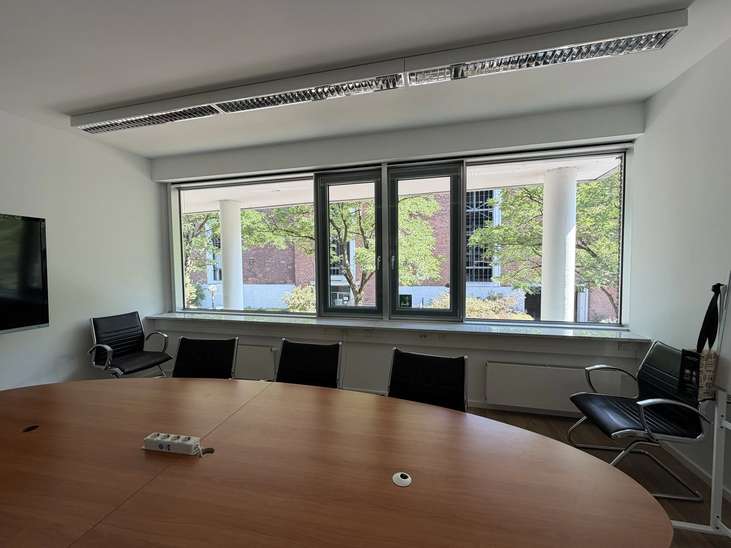 Ein Besprechungszimmer mit runden Holztischen, schwarzen Stühlen und großen Fenstern, durch die man Bäume und ein Gebäude sieht.